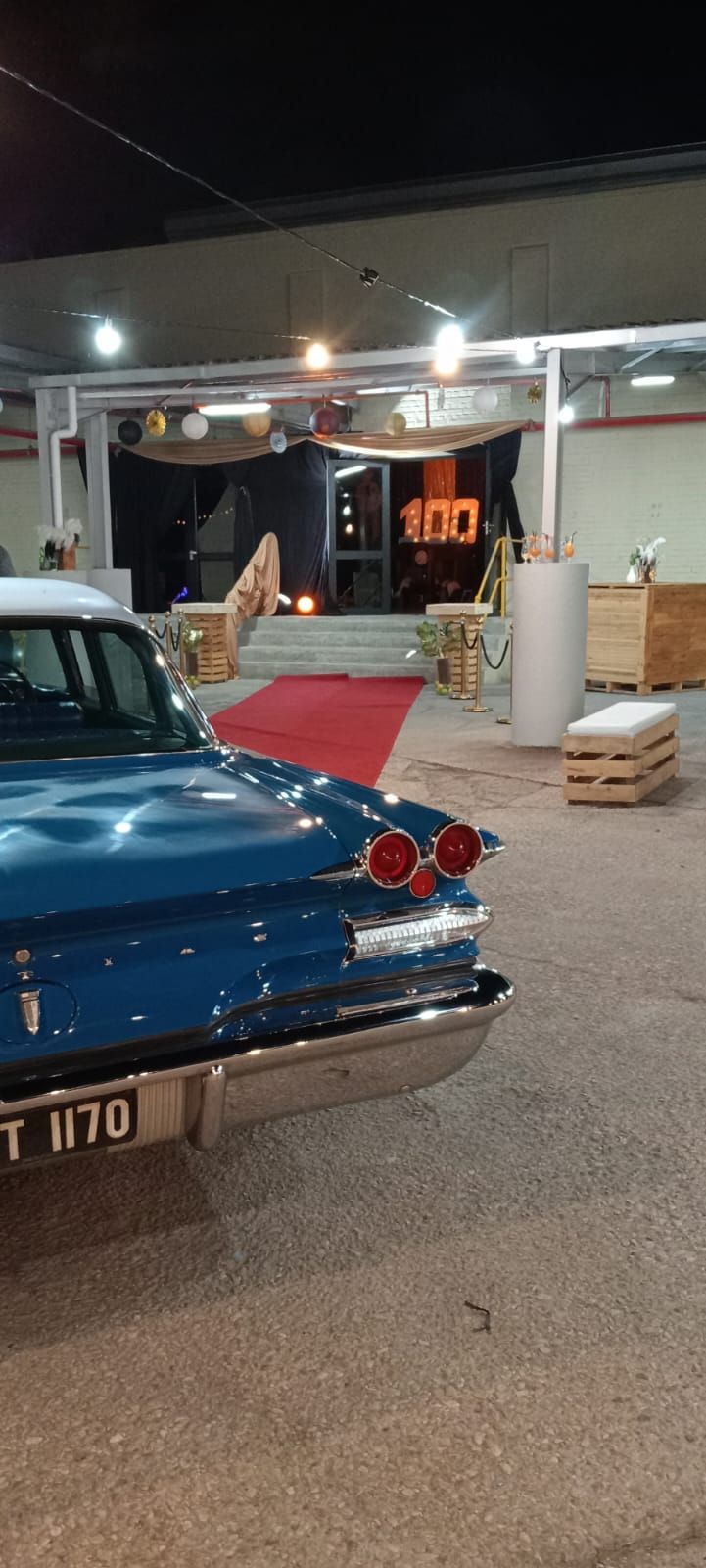 A blue classic car parked near a building entrance with a red carpet. The number 