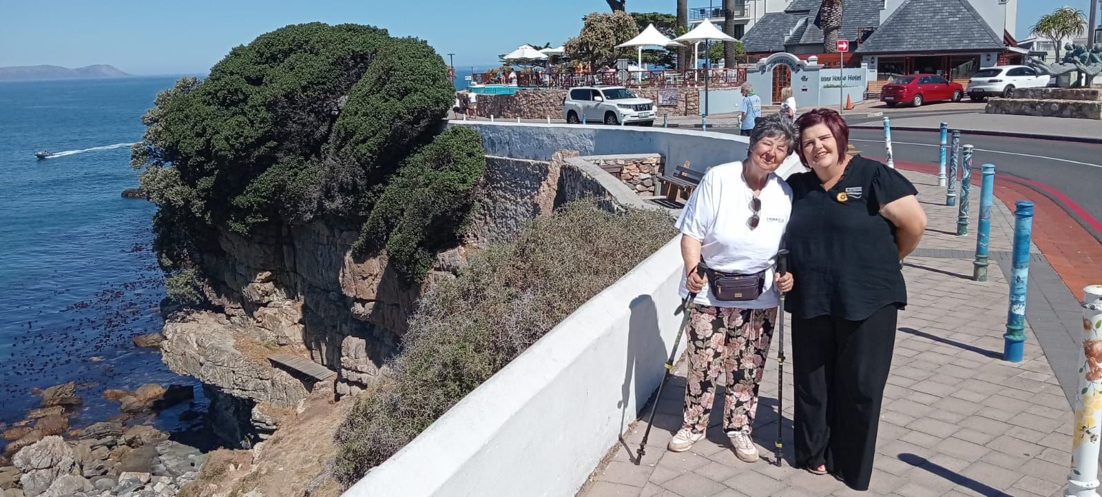 Two women posing on a seaside walkway. One uses a walker. Ocean and buildings in the background.