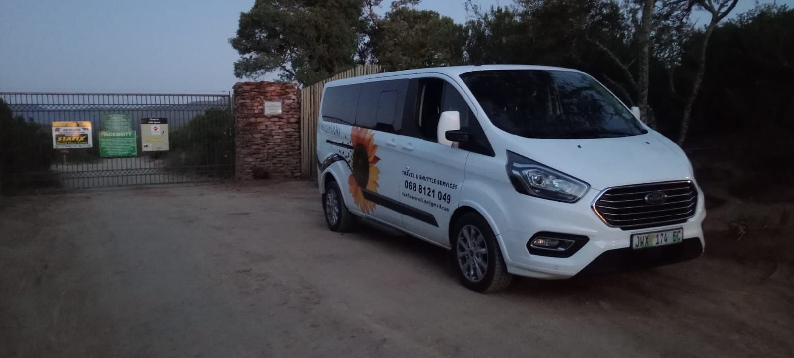 White Ford Transit van parked on a dirt road near an entrance with signs and closed gate.