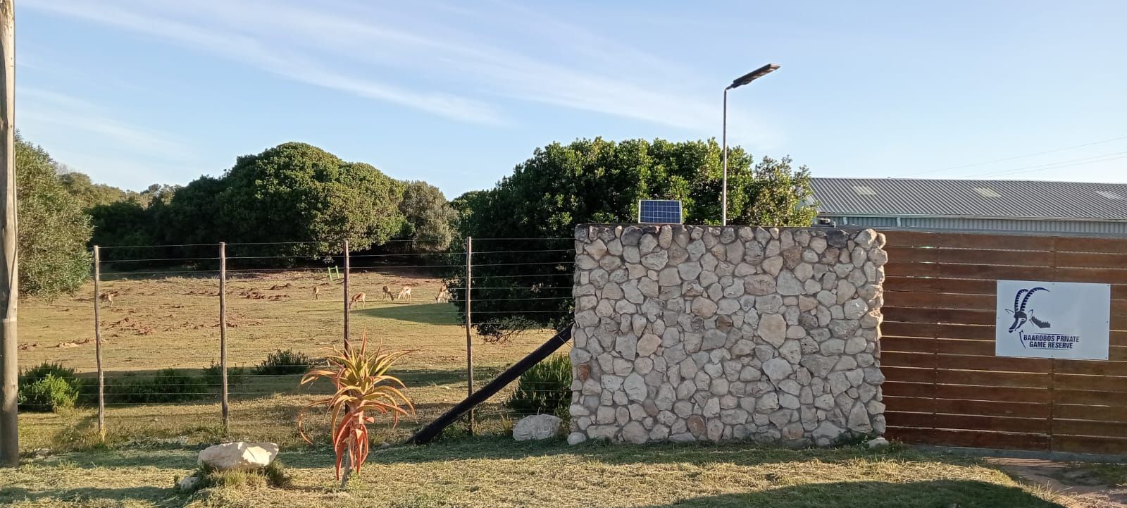 A stone and wooden wall with a sign in front of trees and a field under a blue sky.