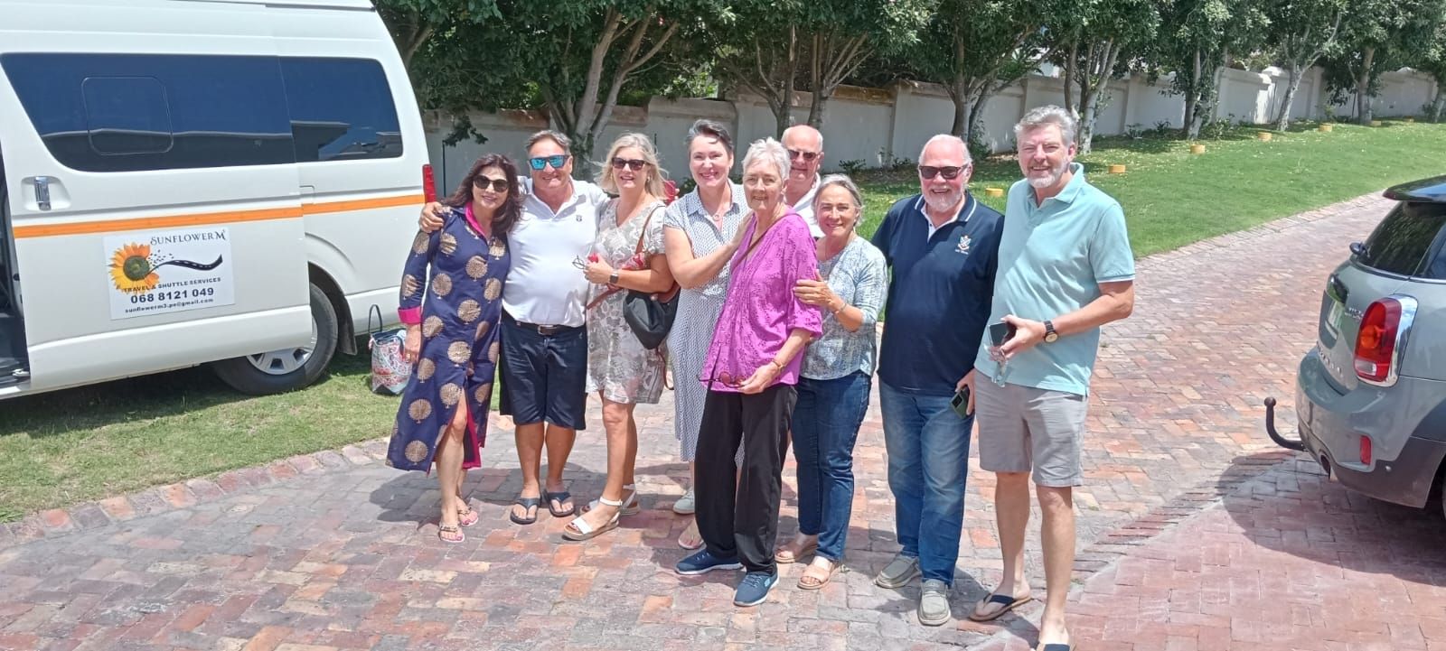 Group of people posing by a white van, on a paved area, trees in background.