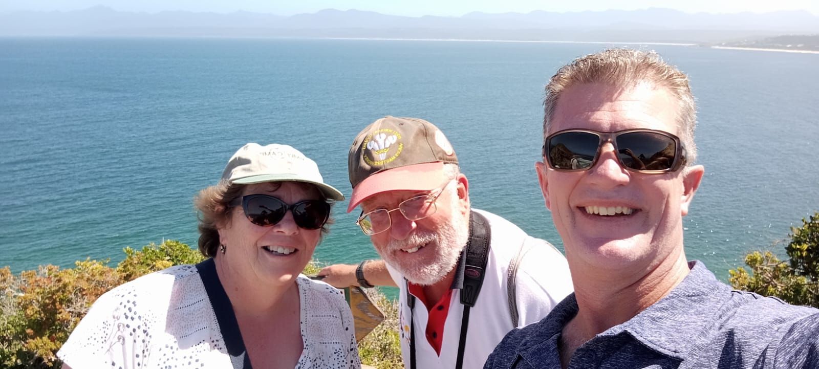 Three people smiling for a selfie overlooking the ocean and mountains. Sunny day.