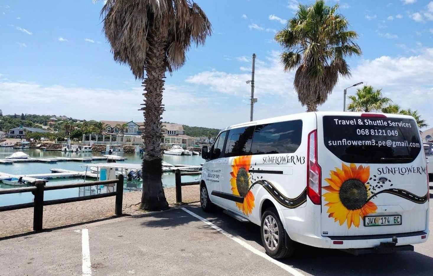 White van with sunflower logo parked by a marina, palm trees, blue sky.