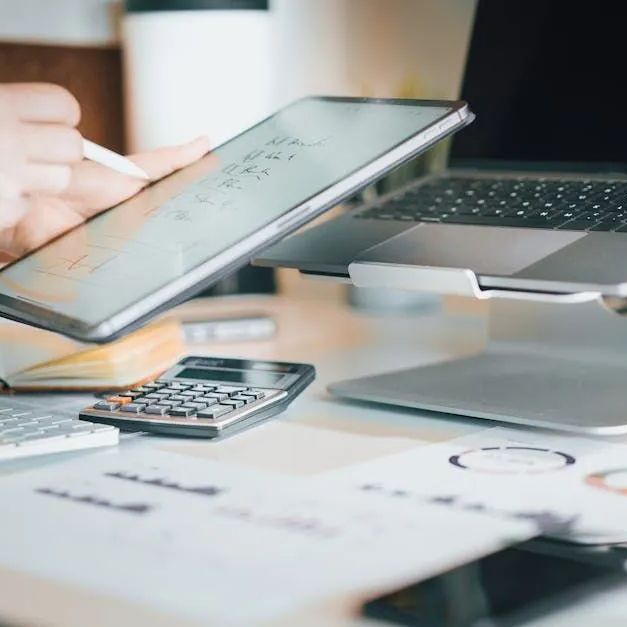 A person is using a tablet computer on a desk next to a laptop and calculator.