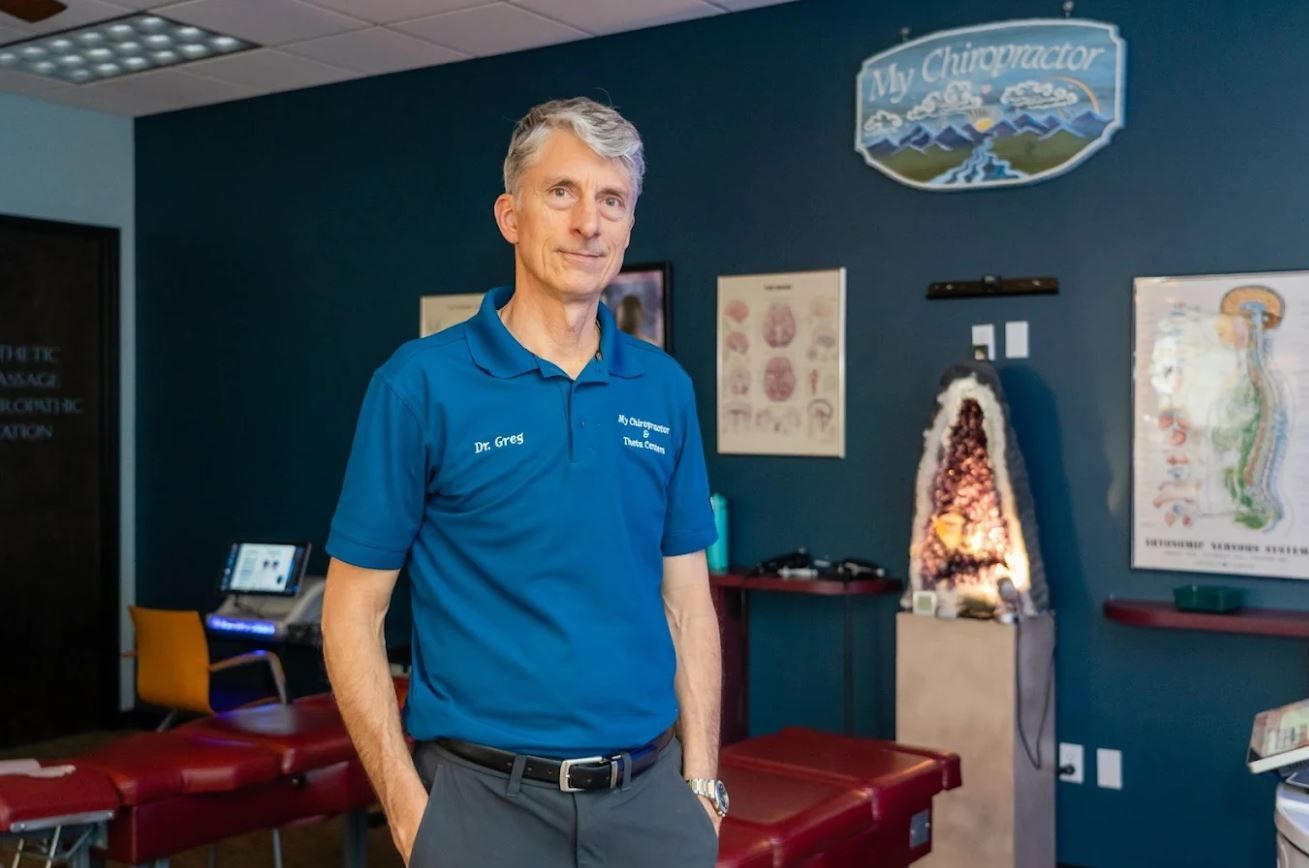 Chiropractor in blue shirt stands in his office; charts on wall, examining table visible.