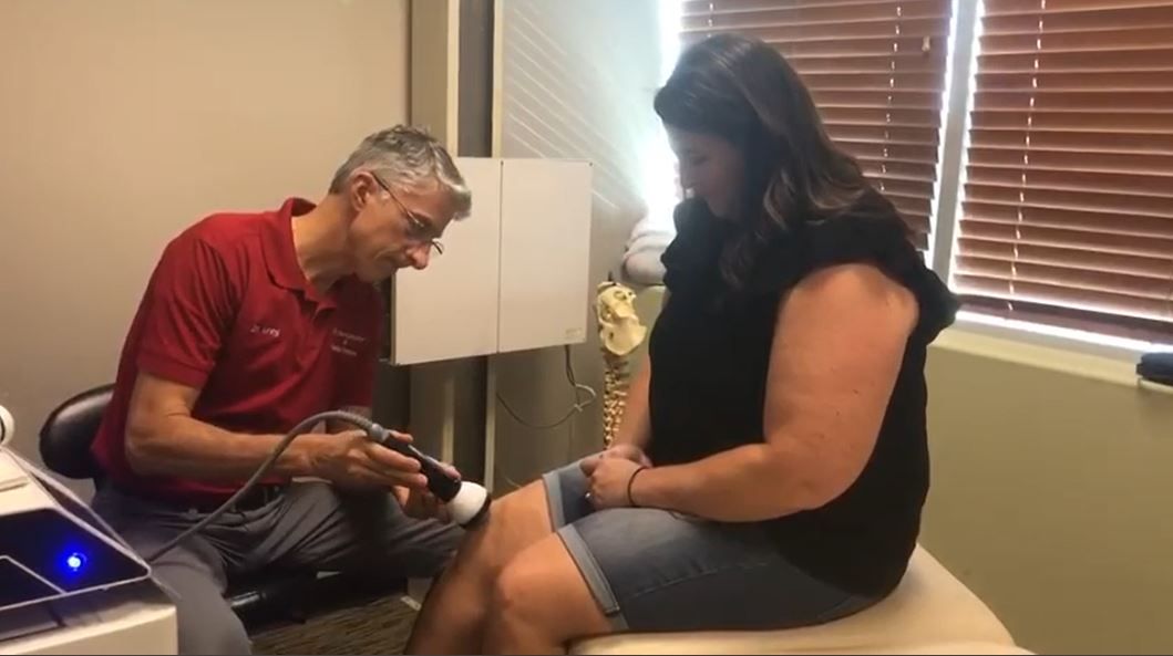Doctor treating patient's knee with a device in an office. Doctor in red shirt, patient in black top, window in background.