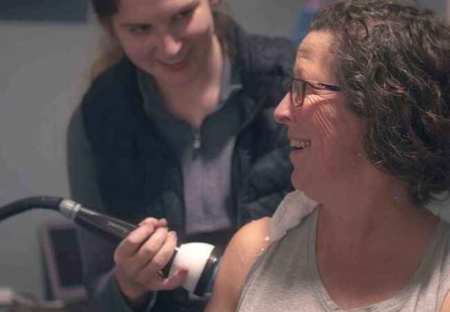Woman receiving shoulder treatment with a medical device; smiling therapist, indoor setting.