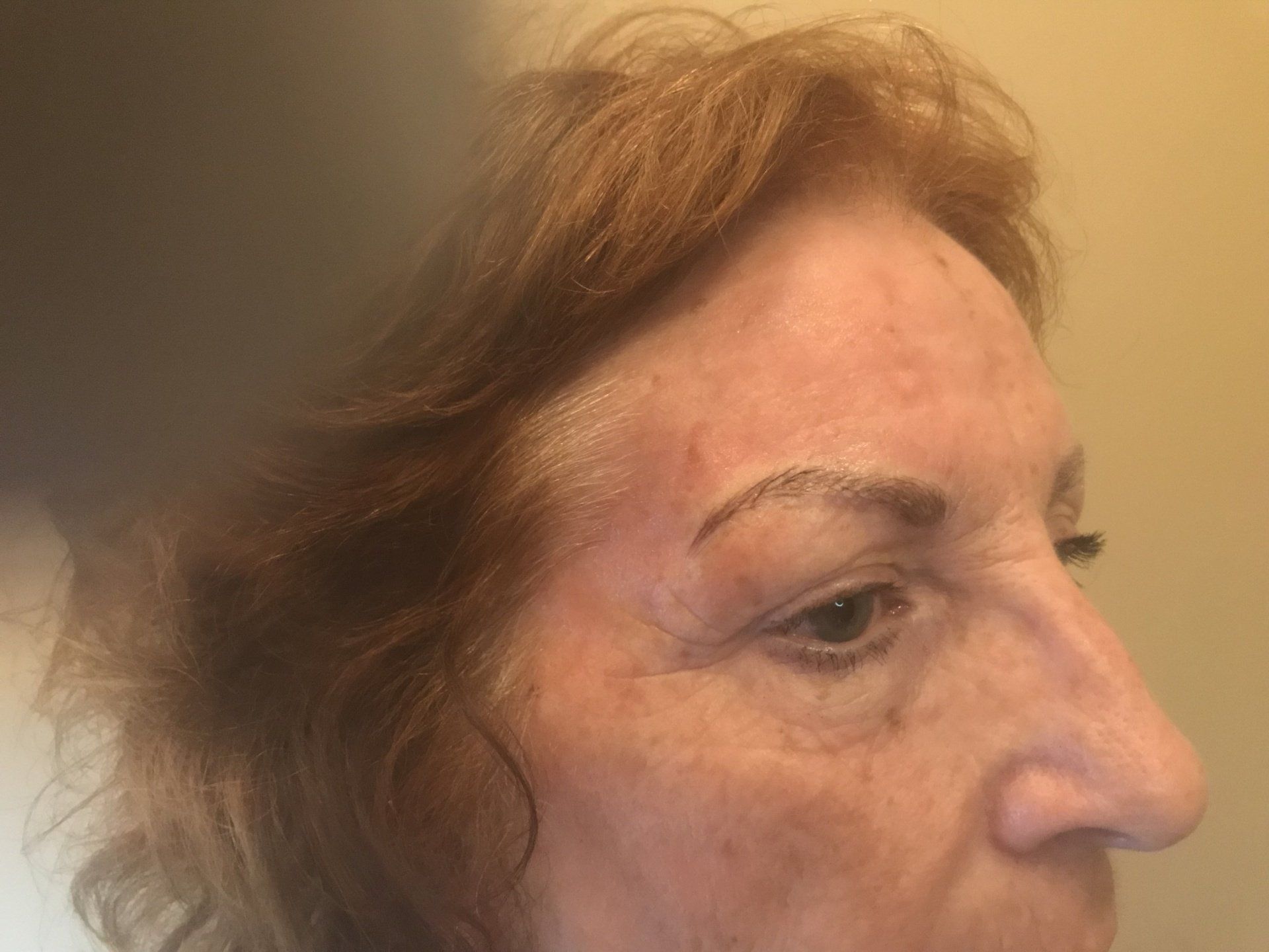 Profile of an older woman with reddish hair and freckles, looking right. She has dark eyebrows.