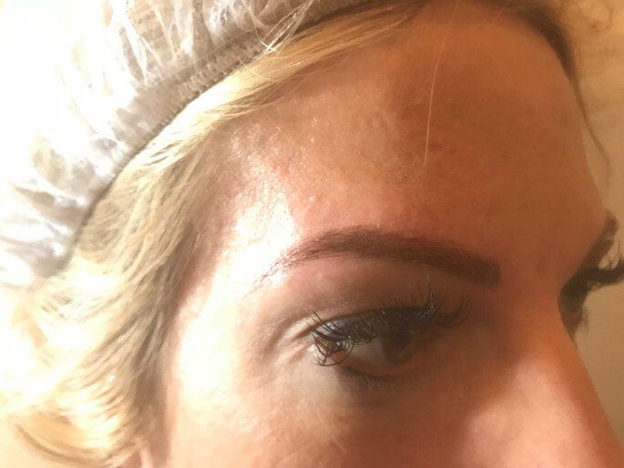 Woman's eye and eyebrow, the brow is colored and shaped, blond hair and a head covering visible.