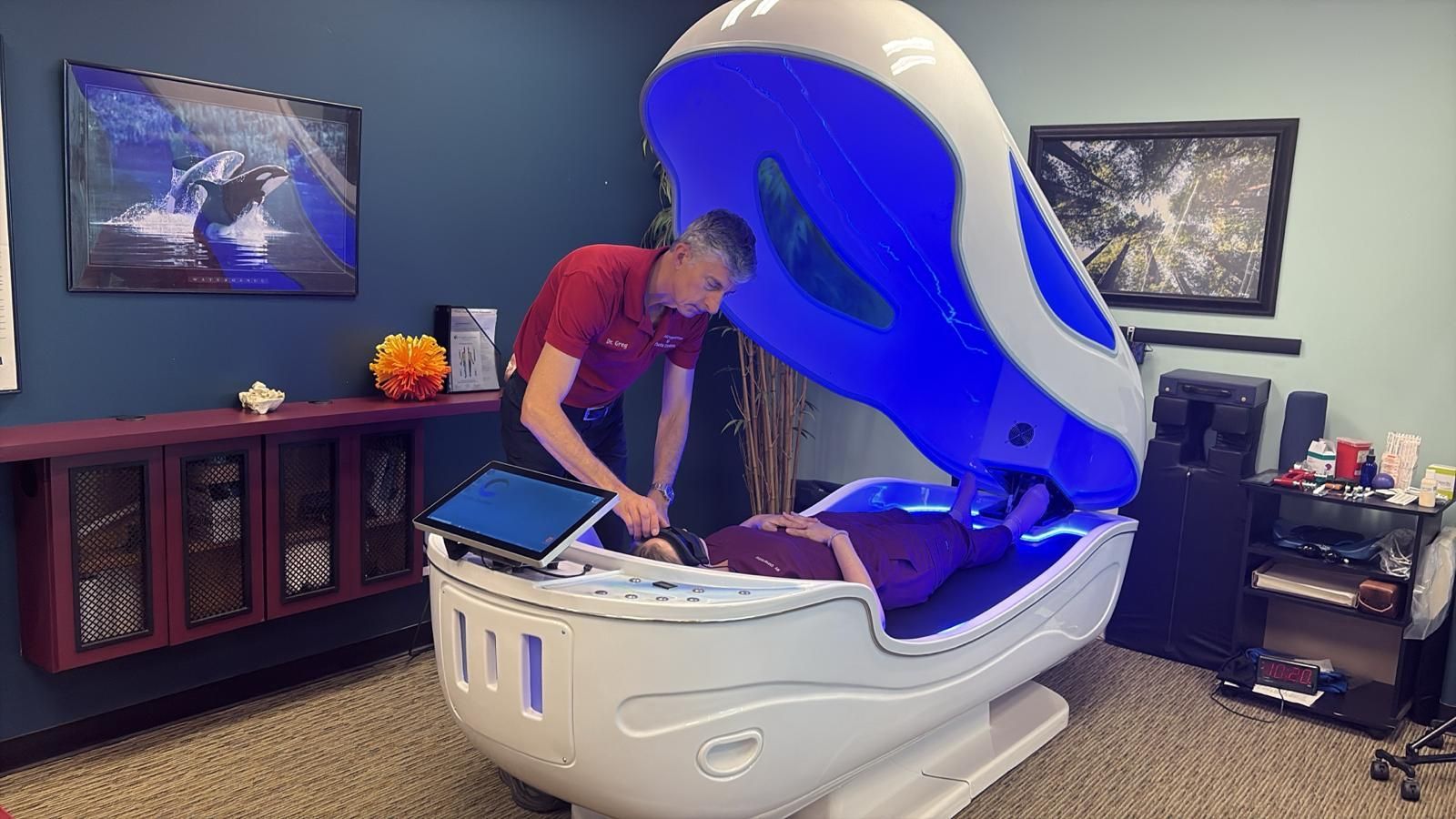 Man assisting another person in a hydrotherapy pod. White and blue pod in a room with a desk and a dark blue wall.