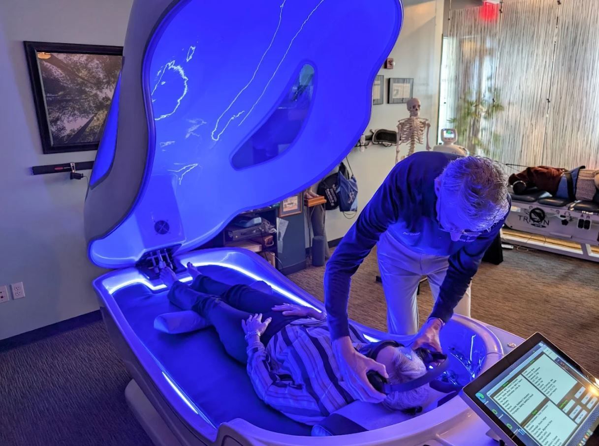 A person lies in a blue light therapy bed, receiving assistance from another person in a brightly lit room.