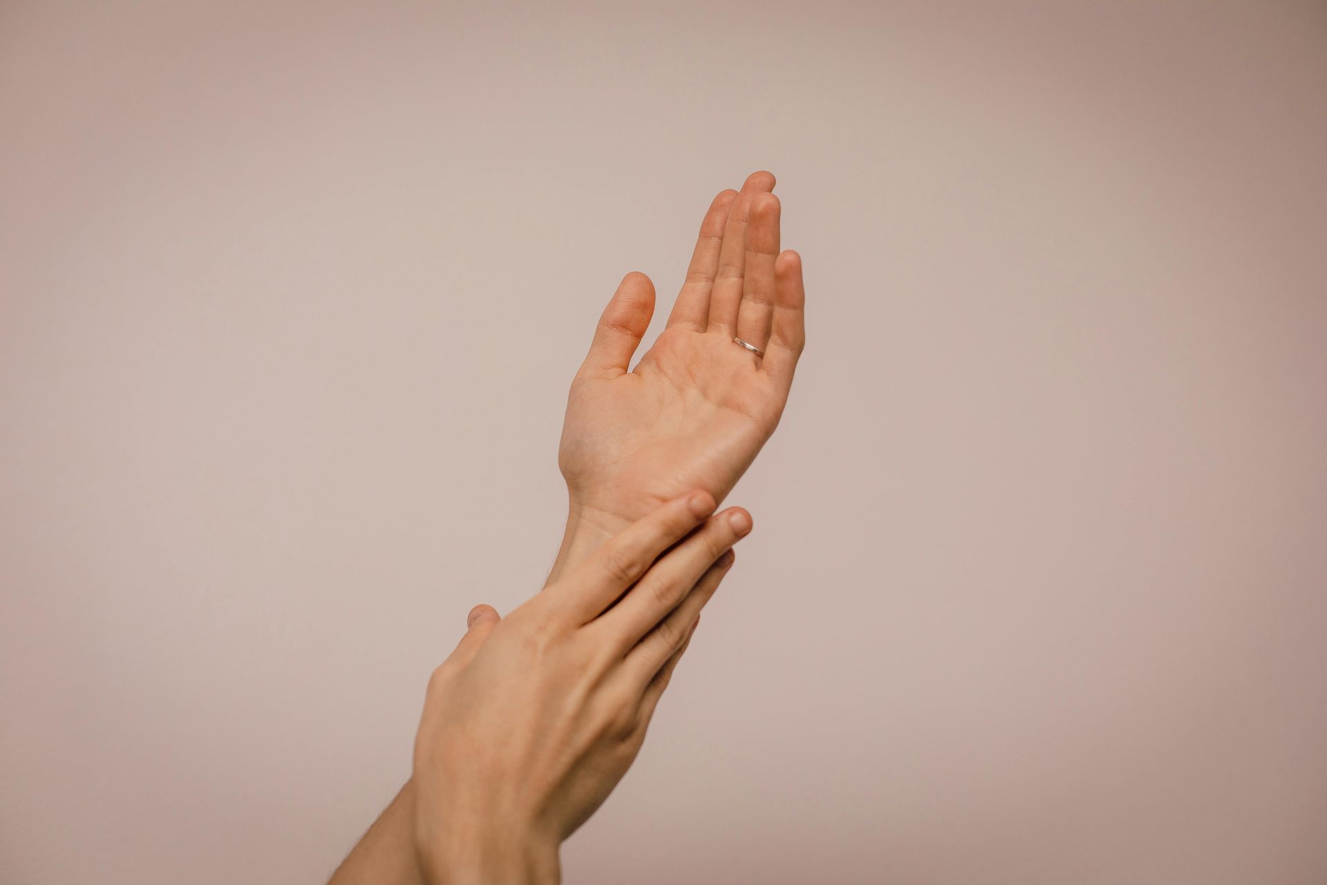 A person's hands, one palm up, the other touching the wrist, against a pink background.