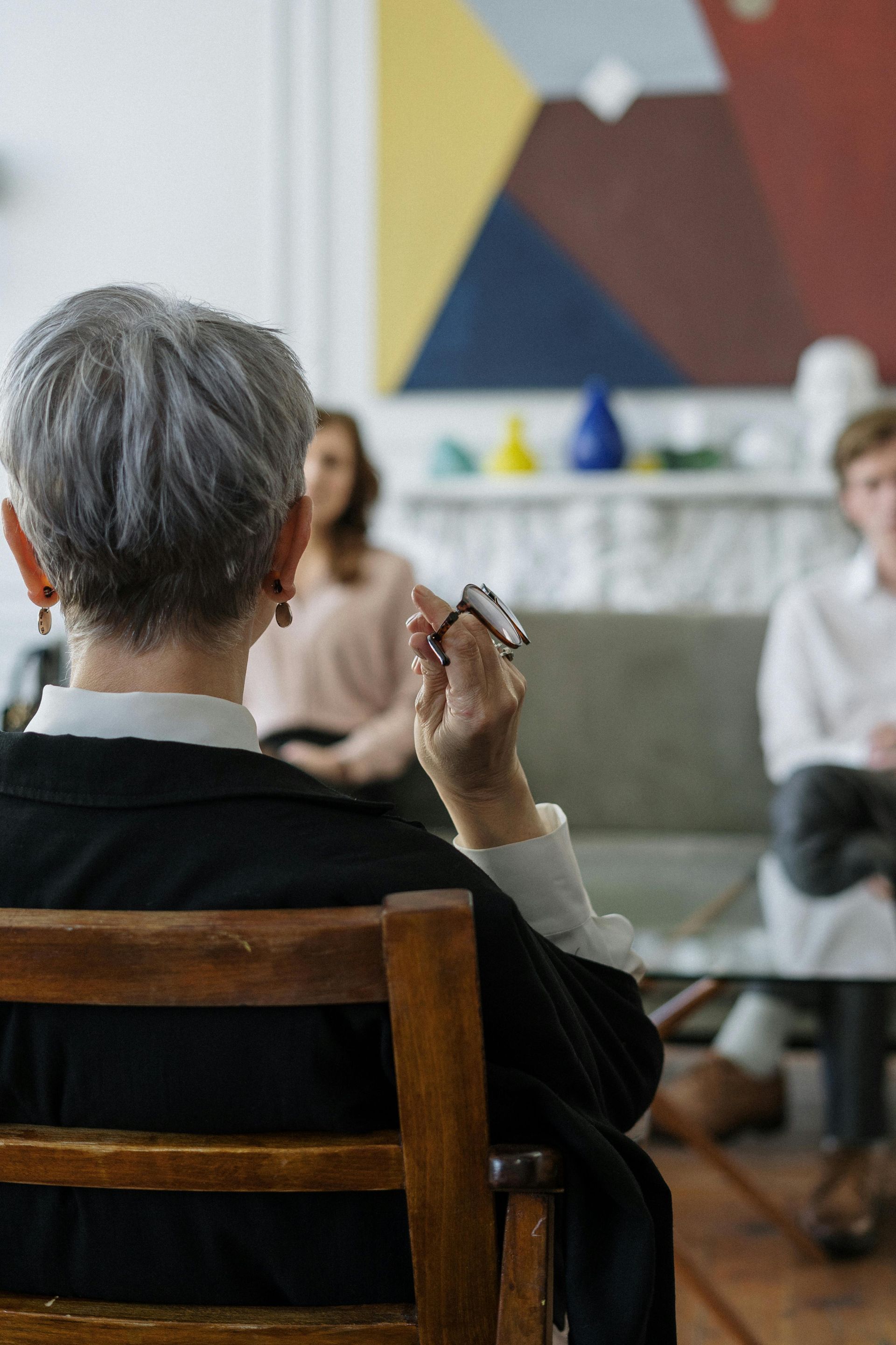 Woman therapist in a session, holding pen, facing two people on a couch. Neutral-toned room, abstract art.