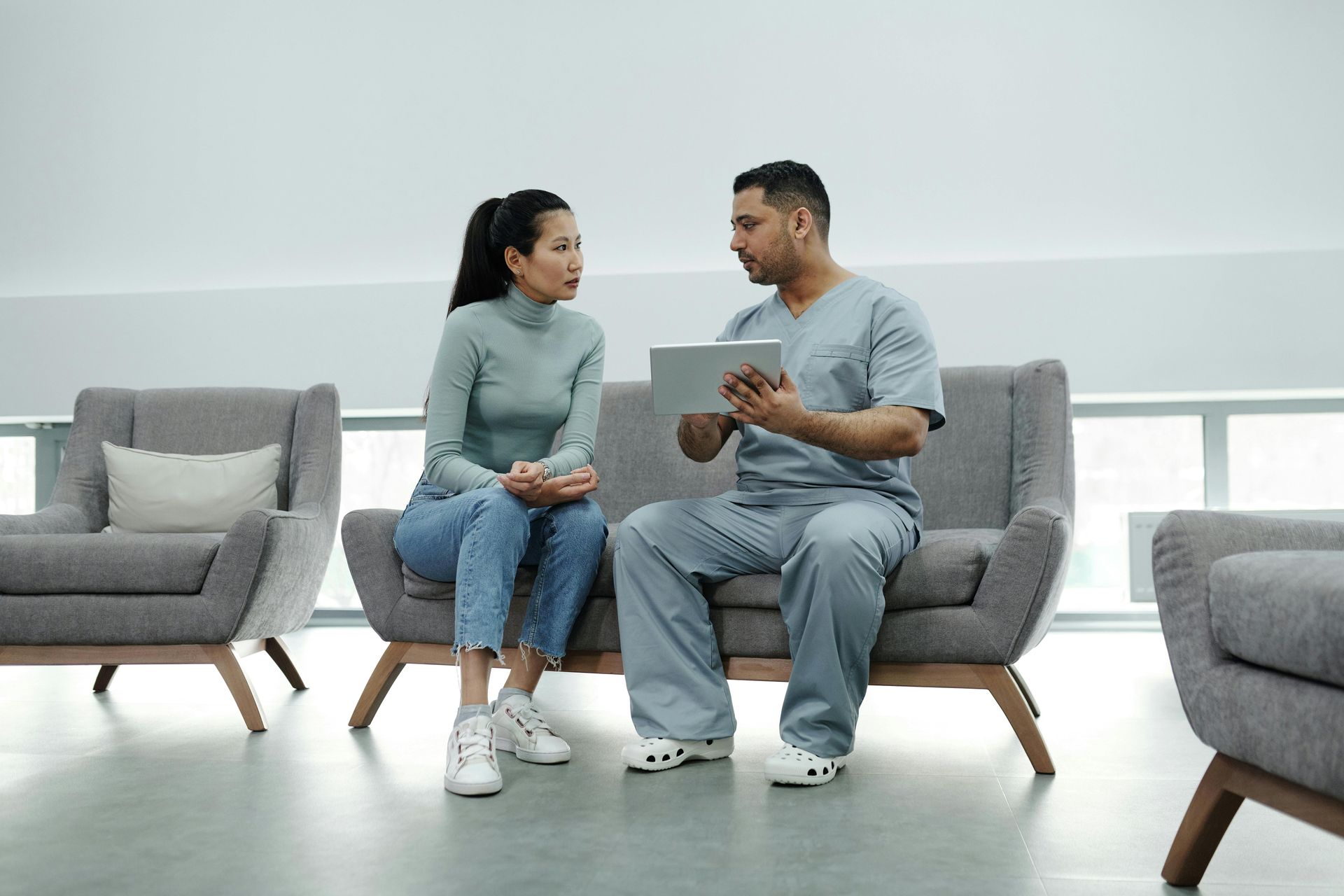 A doctor in scrubs explains something on a tablet to a seated patient in a modern waiting room.
