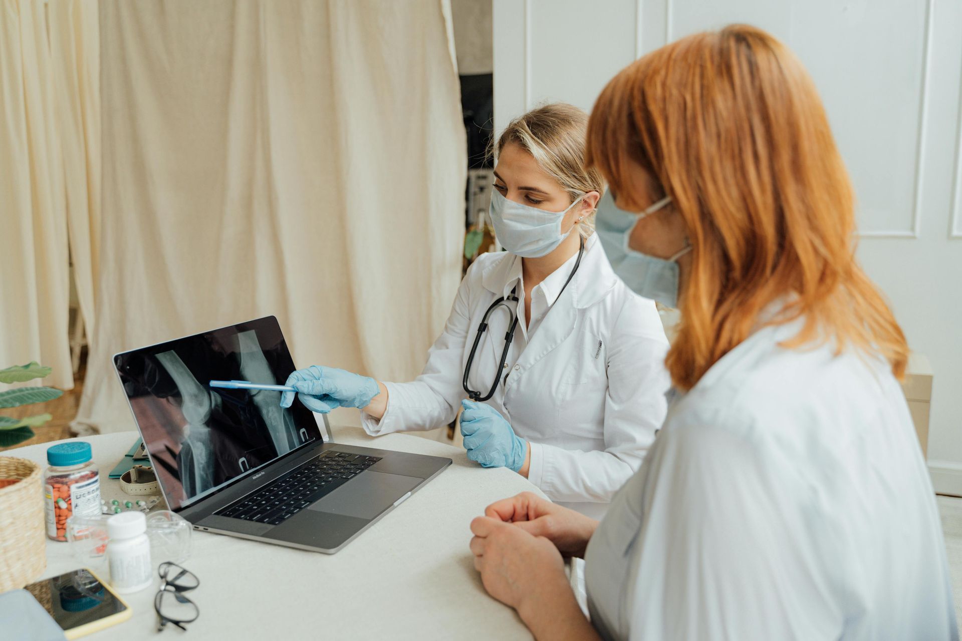 Doctor in mask points at knee X-ray on laptop screen for a red-haired patient, both in medical setting.