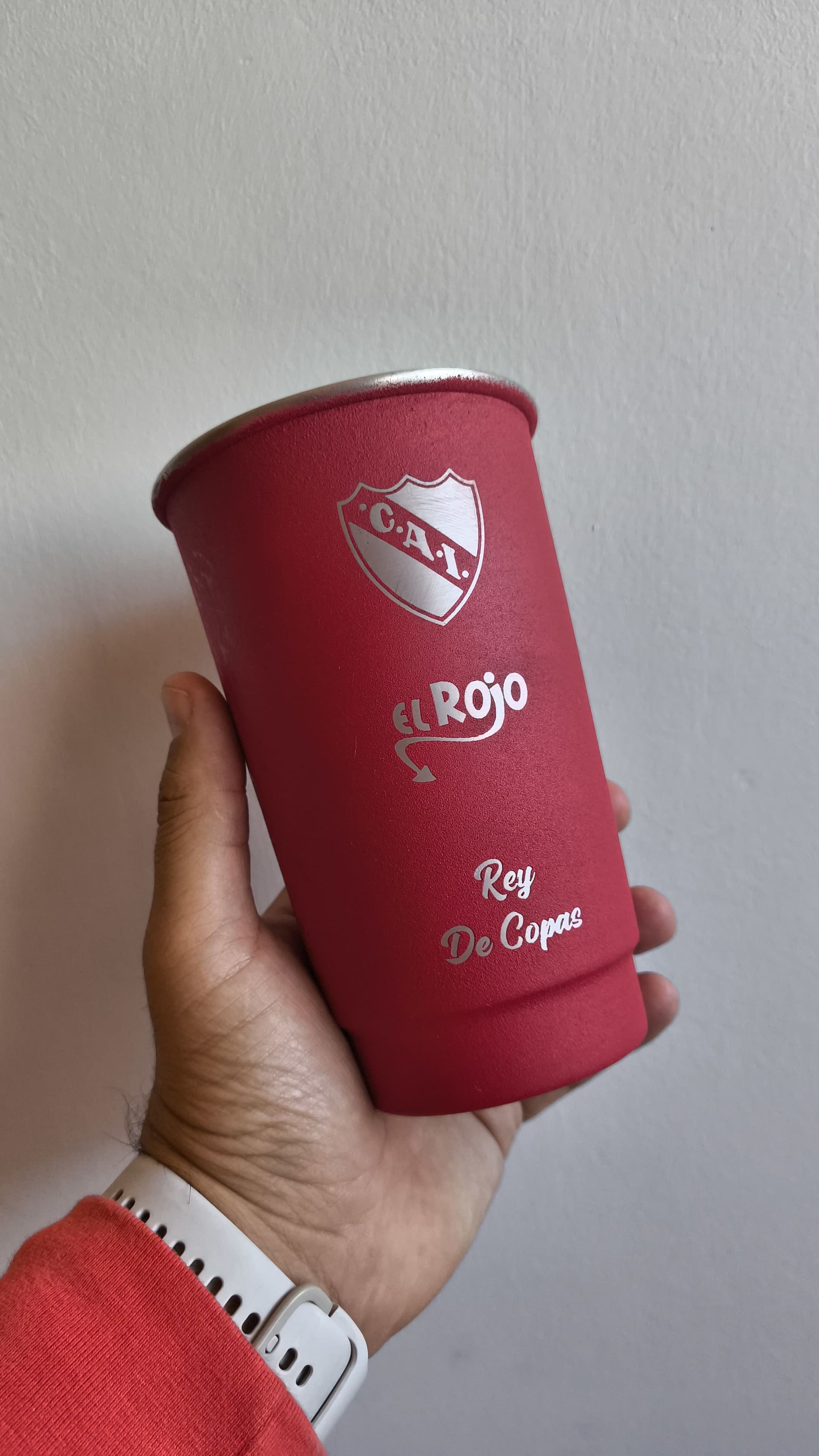 Persona sosteniendo una taza roja con el logo de "El Rojo" contra una pared blanca.