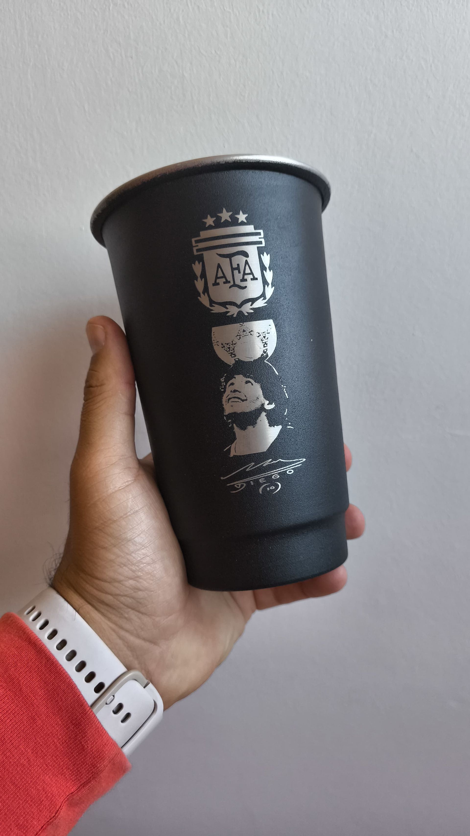 Una mano sosteniendo un vaso térmico negro con el emblema del equipo