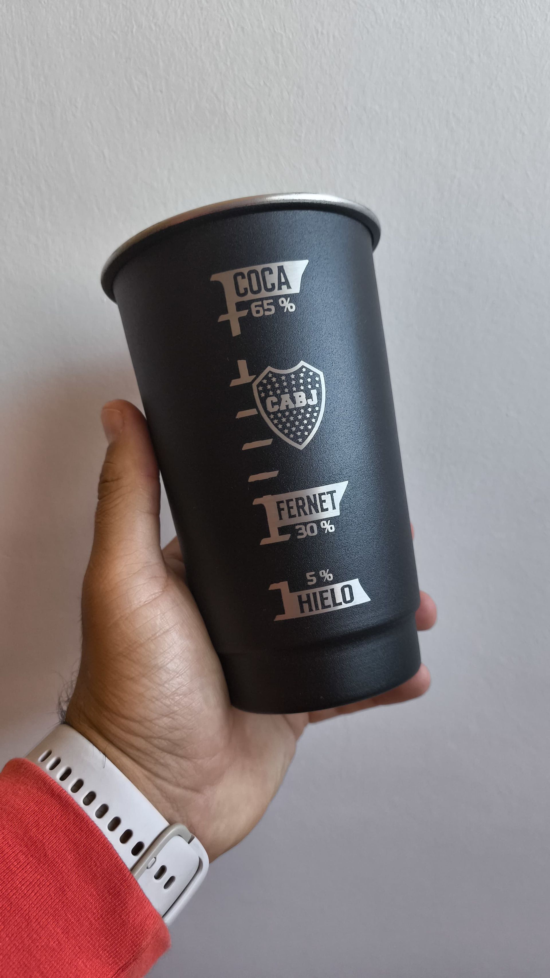 Mano sosteniendo una taza de metal negro con marcas grabadas y el logo de Boca Juniors.