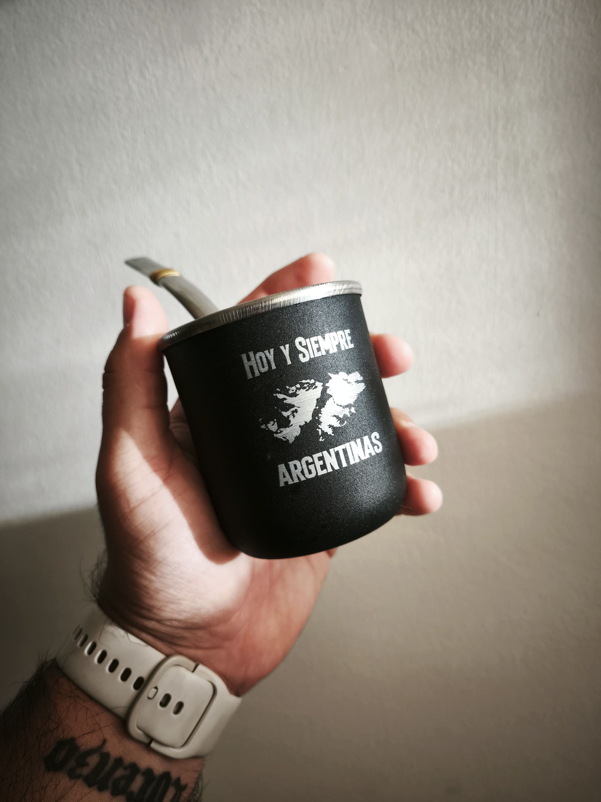 Mano sosteniendo un mate negro con una pajita plateada. El mate tiene la bandera argentina y el texto "Hoy y Siempre Argentinas".