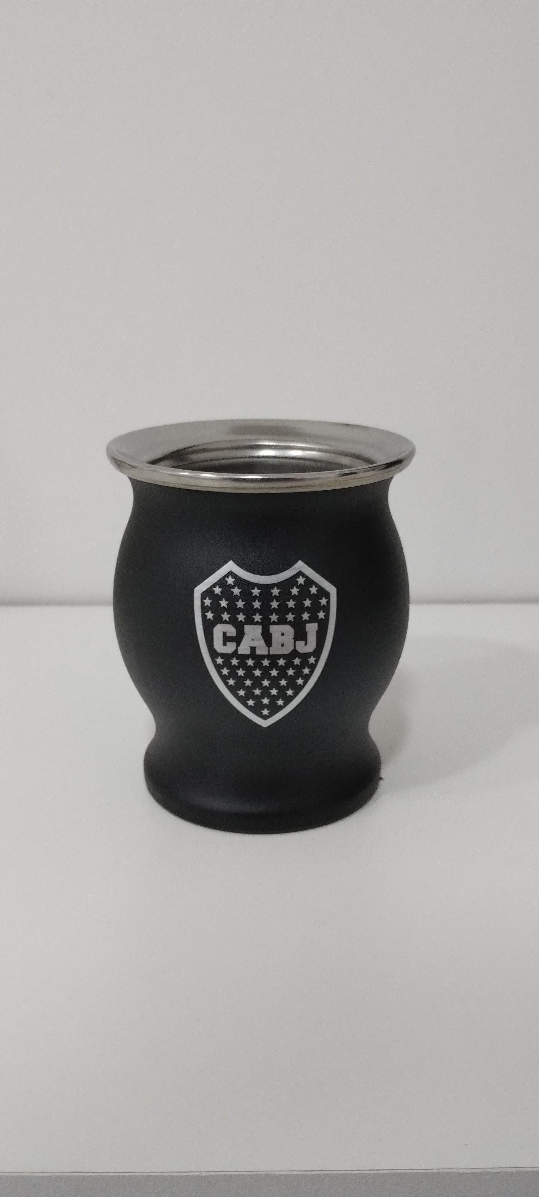 Un mate negro con borde plateado y el logo “CABJ”, colocado sobre una superficie blanca sobre un fondo liso.