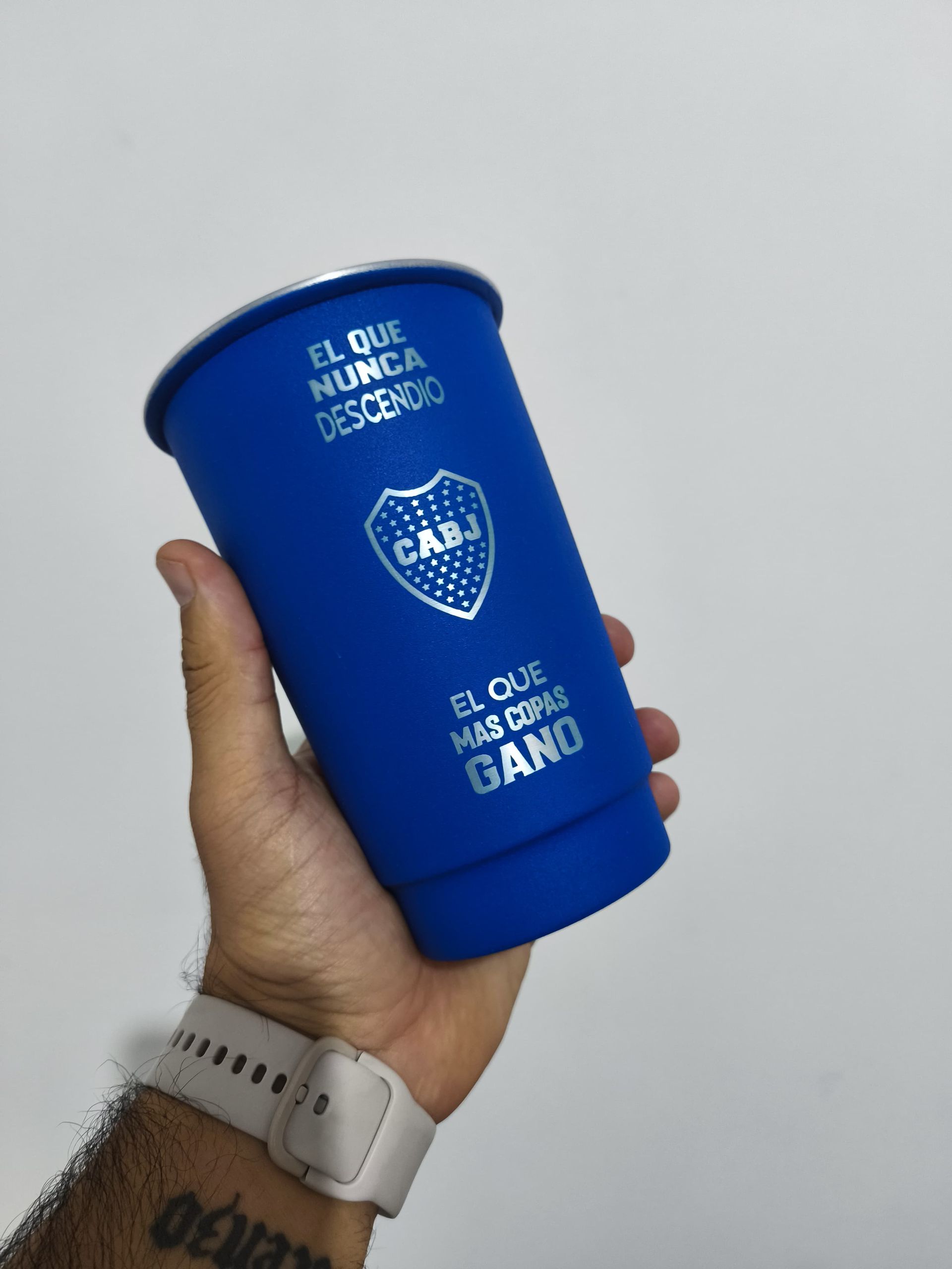 A hand holds a blue Boca Juniors fan cup. The cup has the team logo and text "El que nunca desciende, el que más copas ganó" in white.
