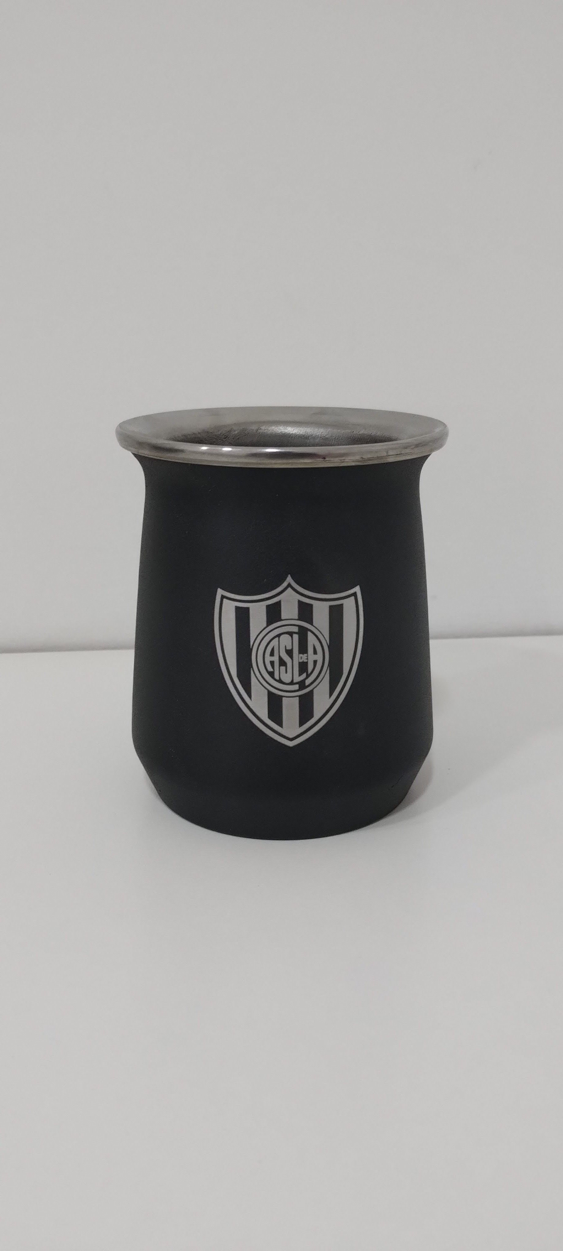 Mate negro con borde plateado y el escudo del Atlético Mineiro. Reposa sobre una superficie blanca contra una pared blanca.