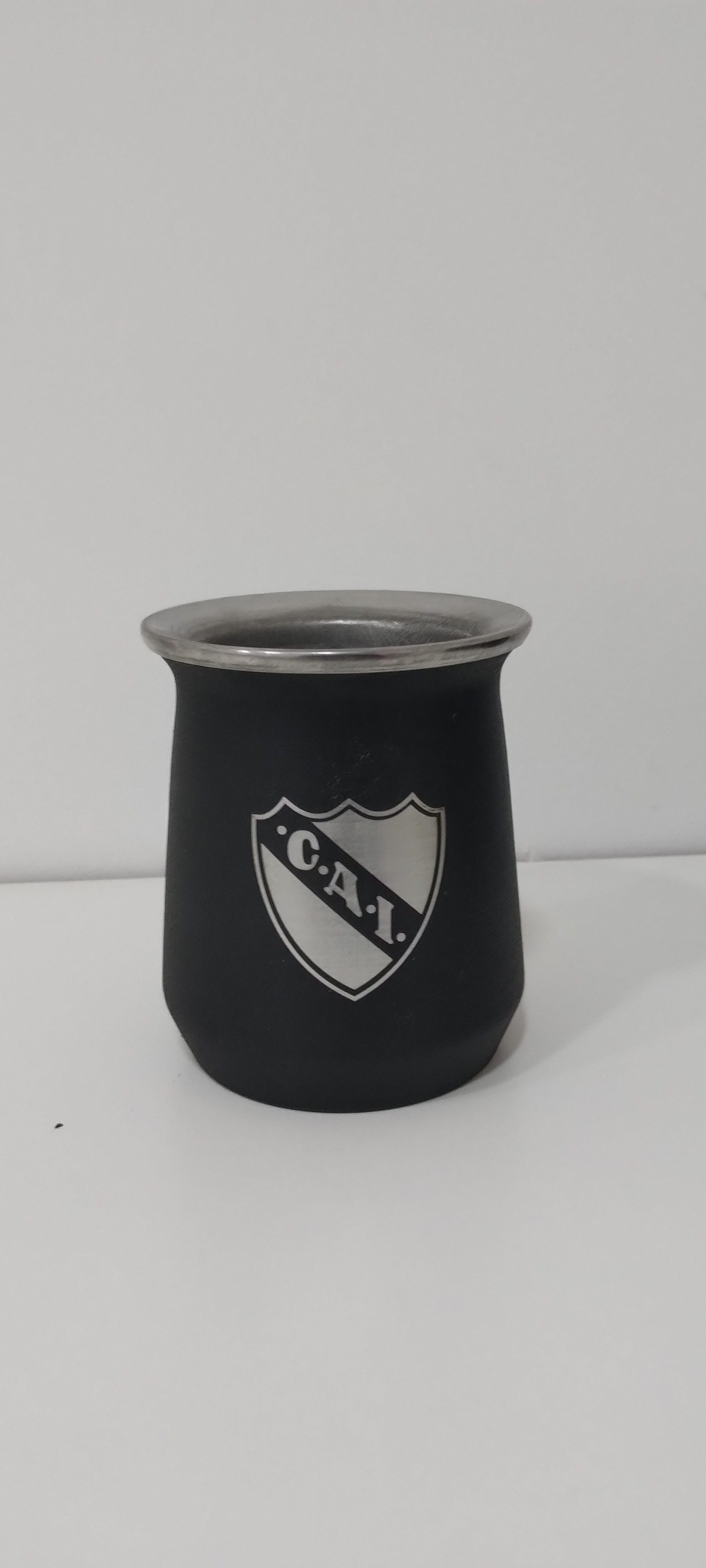 Una calabaza mate negra con borde plateado y un escudo con las letras "CAI". La calabaza está sobre una superficie blanca.