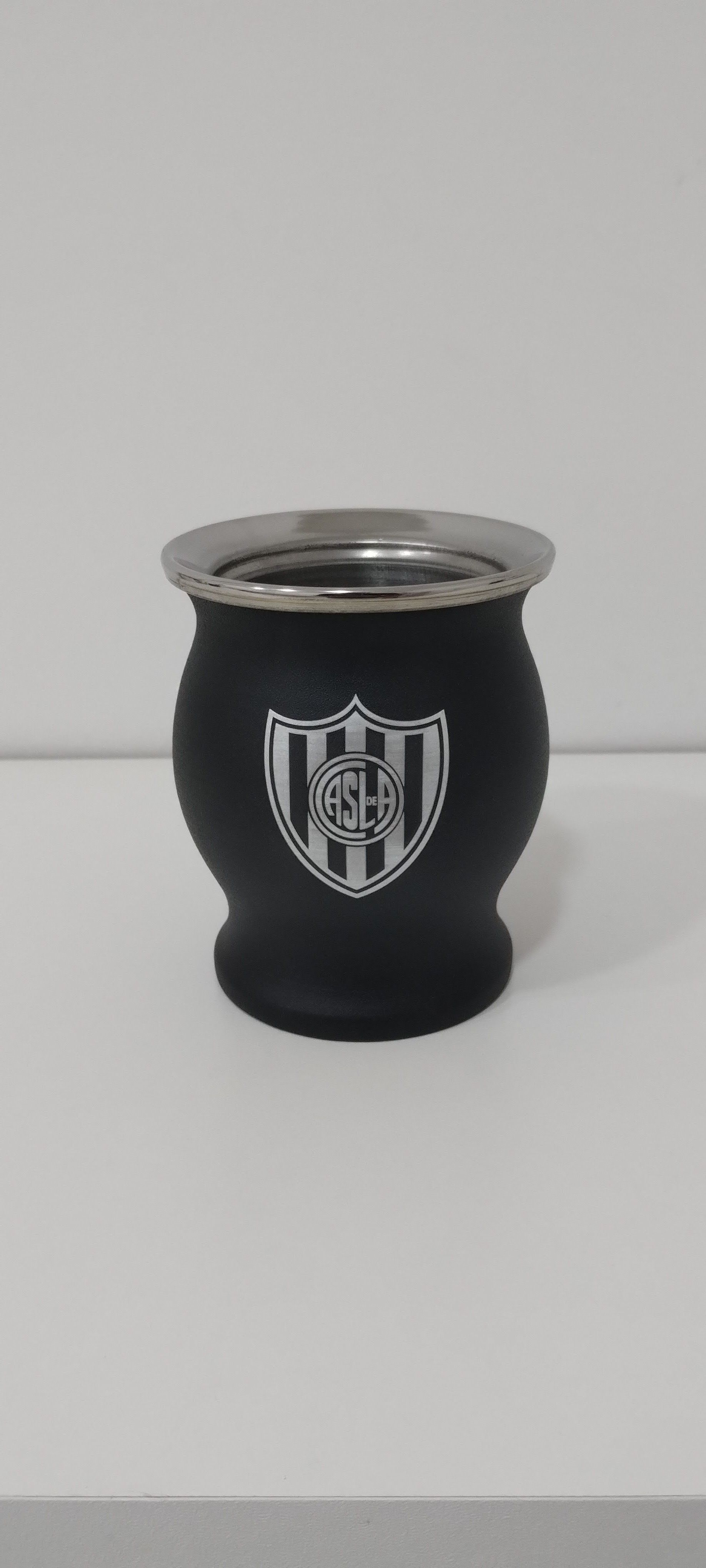 Calabaza negra con borde plateado y el logo del equipo, colocada sobre una superficie blanca.