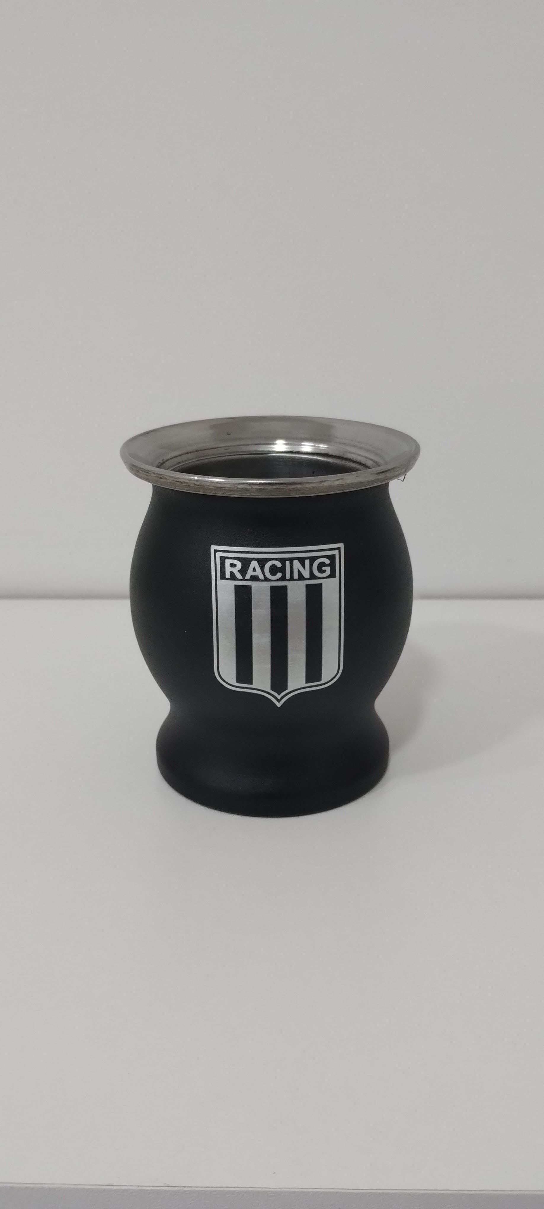 Una calabaza mate negra del Racing Club con borde metálico reposa sobre una superficie blanca. El escudo del equipo está grabado en la calabaza.