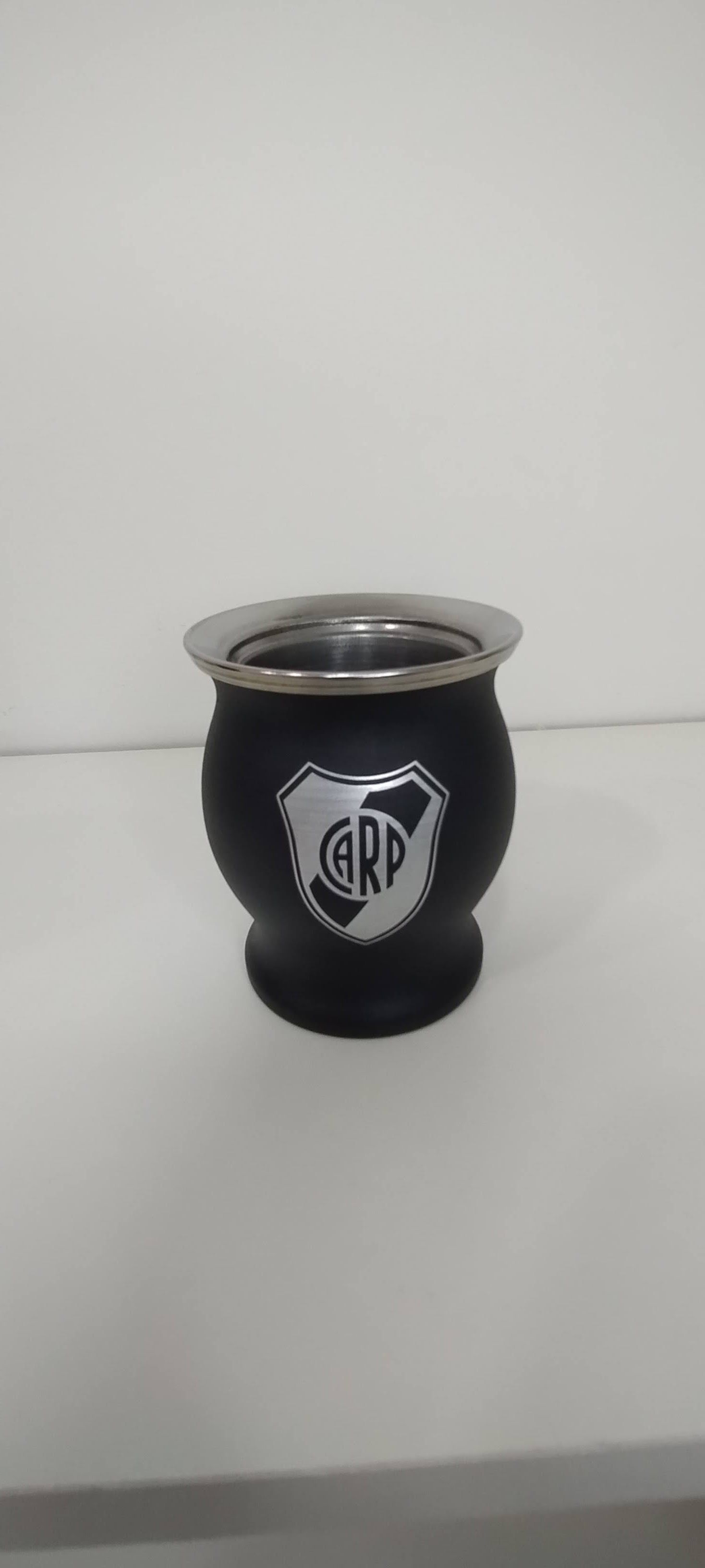 Un mate de color negro con borde plateado y un logo con las letras "CARP" sobre una superficie blanca.