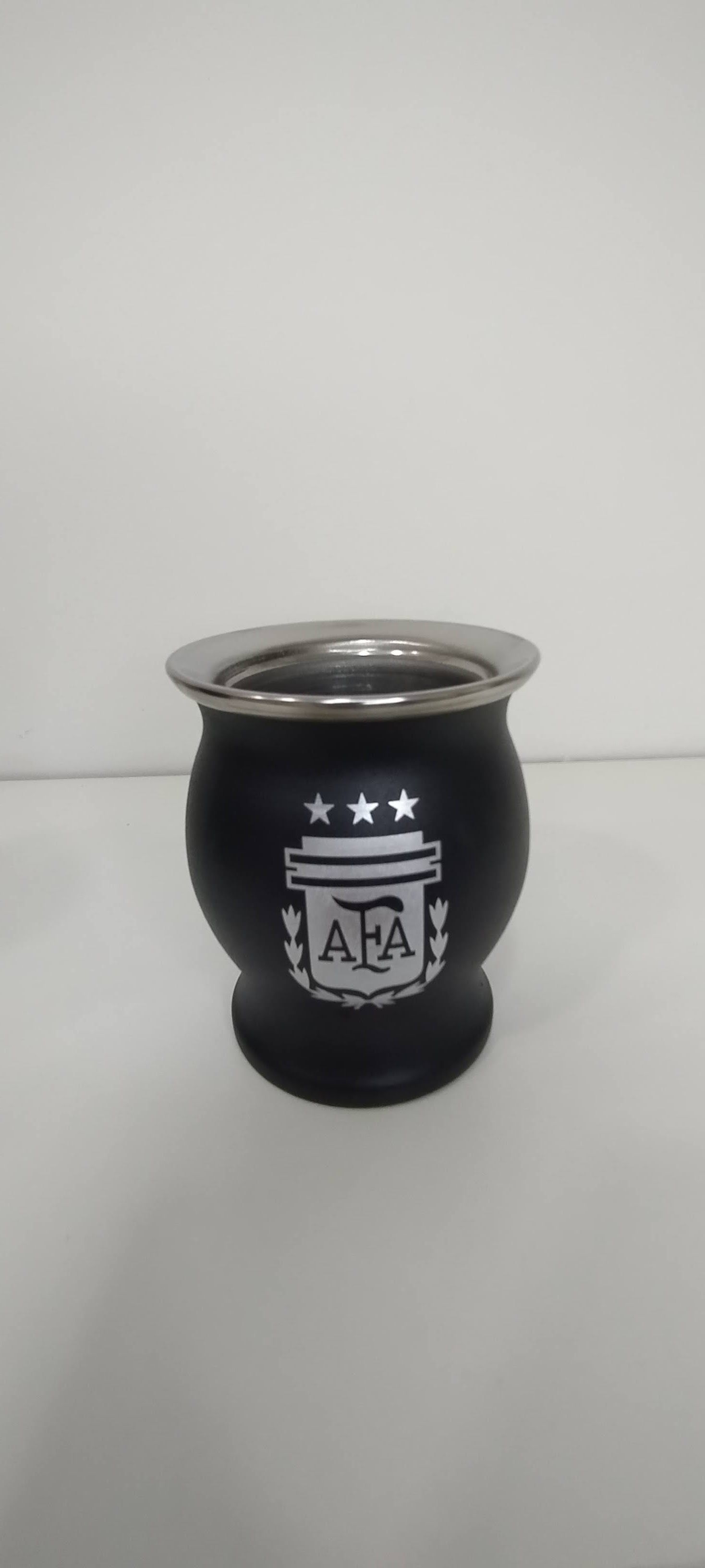 Un mate de color negro con borde plateado y el logo de la selección argentina sobre superficie blanca.