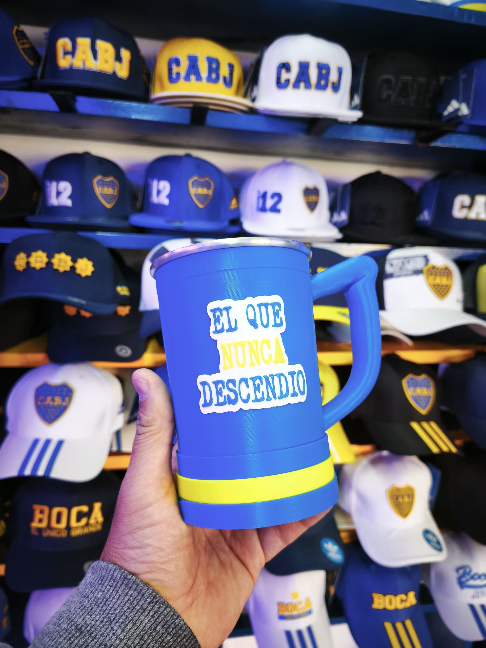 Mano sosteniendo una taza azul de Boca Juniors que dice "El Que Nunca Descendió". Varias gorras de Boca Juniors al fondo.
