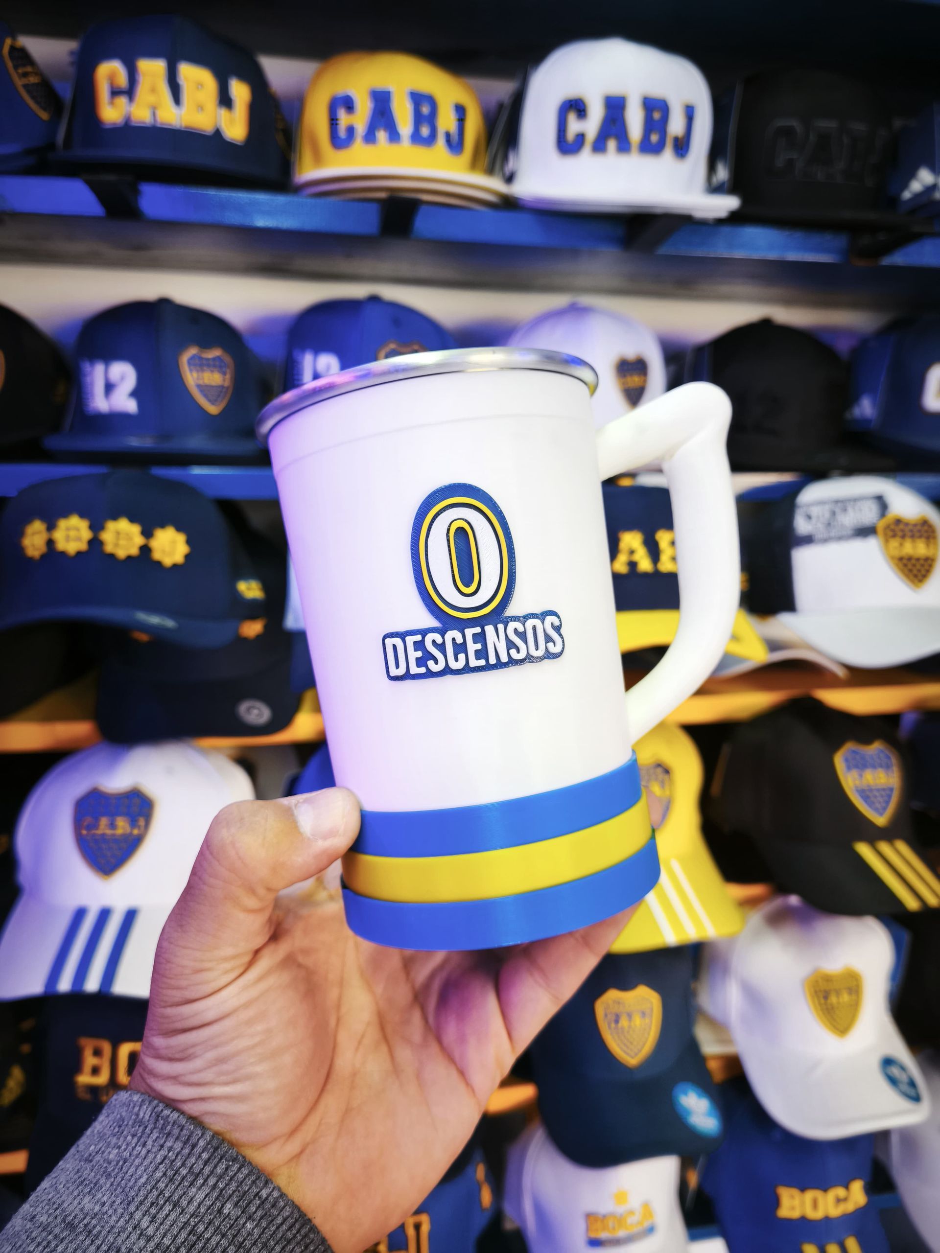 Mano sosteniendo una taza con el logo de Boca Juniors y el texto "Descensos", sobre un fondo de gorras de Boca Juniors.