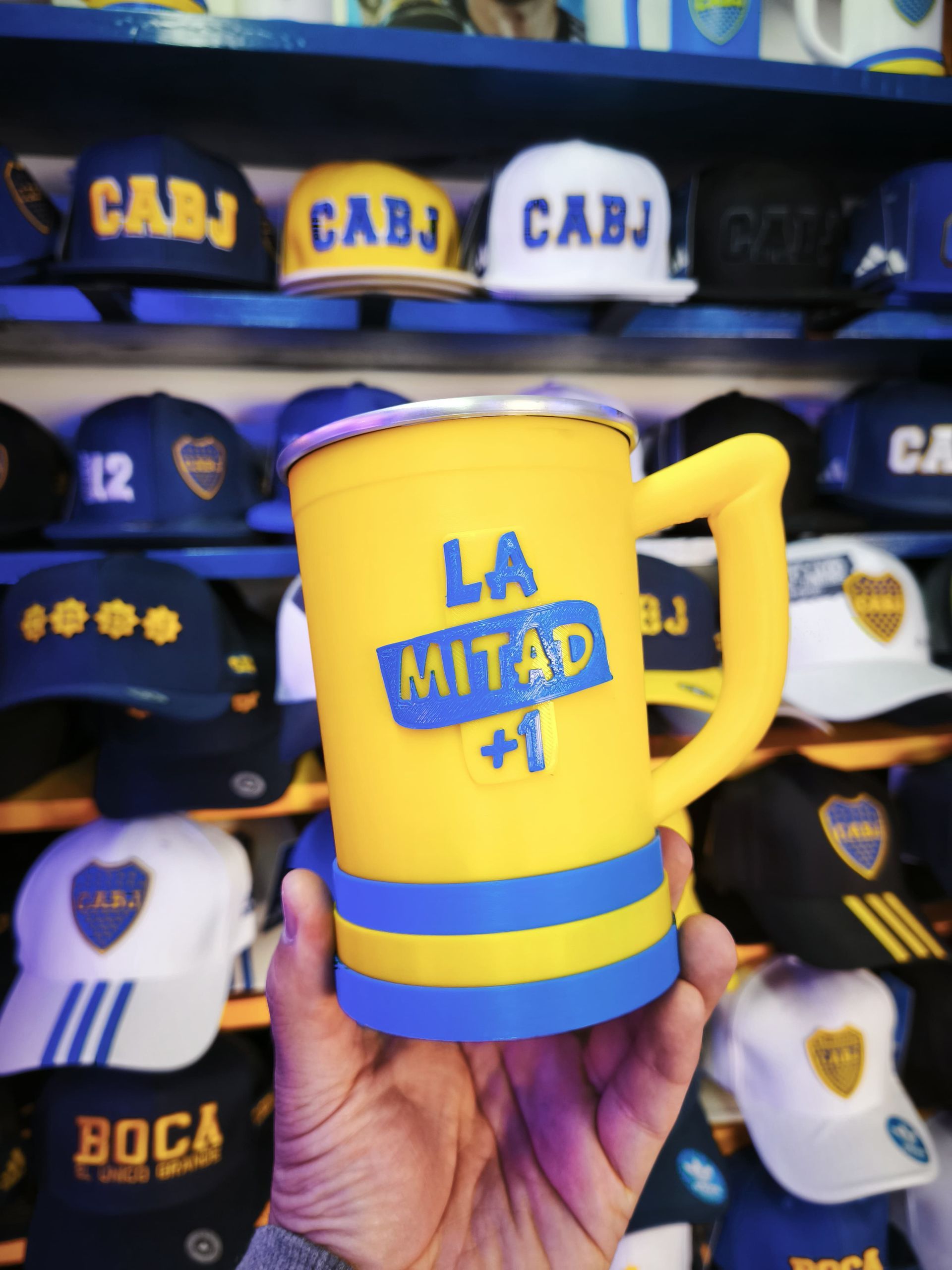 Mano sosteniendo una taza amarilla y azul de Boca Juniors con el texto "LA MITAD +1". Gorras de béisbol con "CABJ" al fondo.