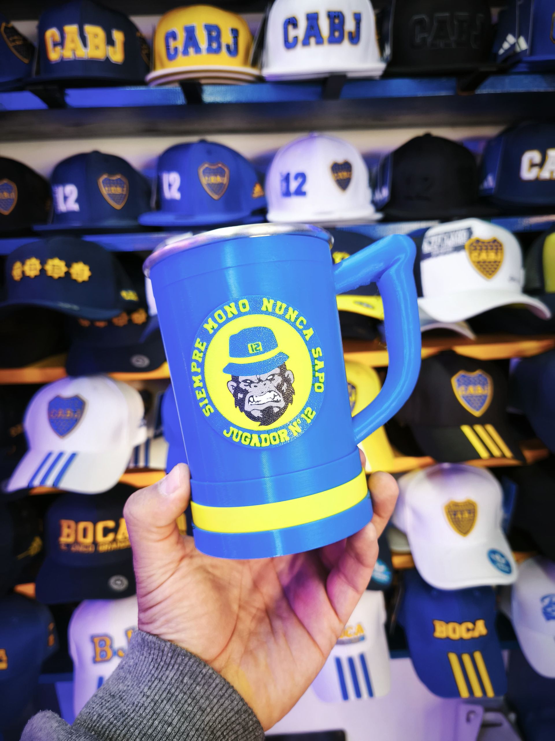 Mano sosteniendo una jarra de cerveza azul y amarilla de Boca Juniors con el logo de un gorila, frente a una pared de gorras de Boca Juniors.