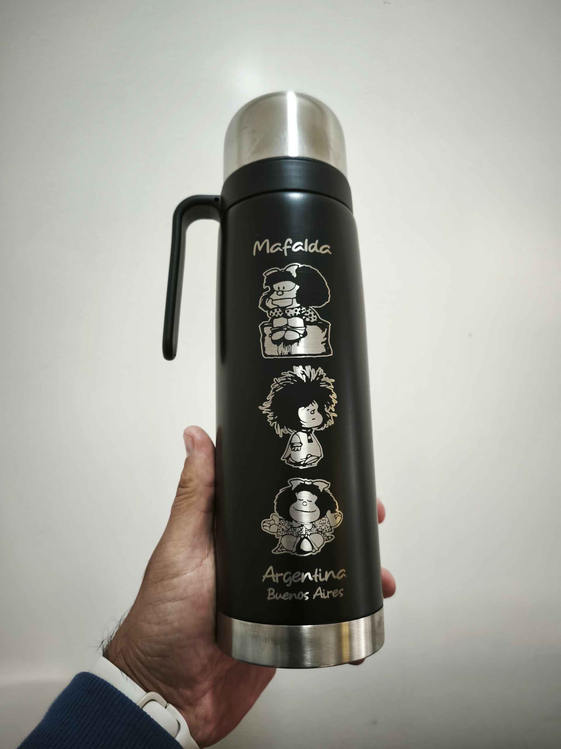 Una persona sostiene un termo negro con grabados de personajes de Mafalda. Tiene tapa, asa y base plateadas.