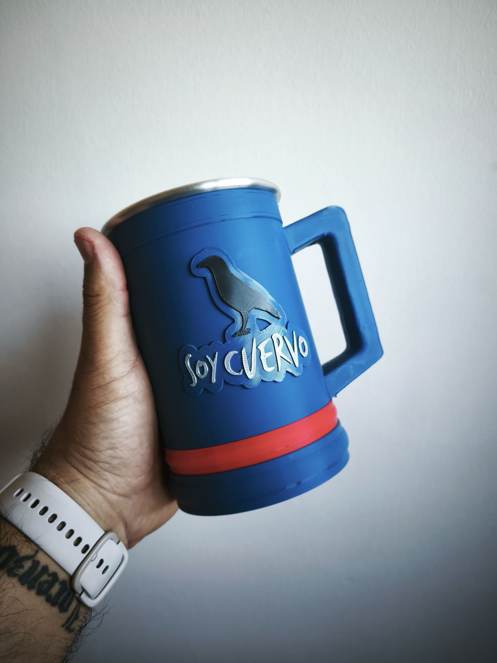 Mano sosteniendo una taza azul con un diseño de cuervo y el texto "Soy Cuervo", banda roja, sobre un fondo blanco.