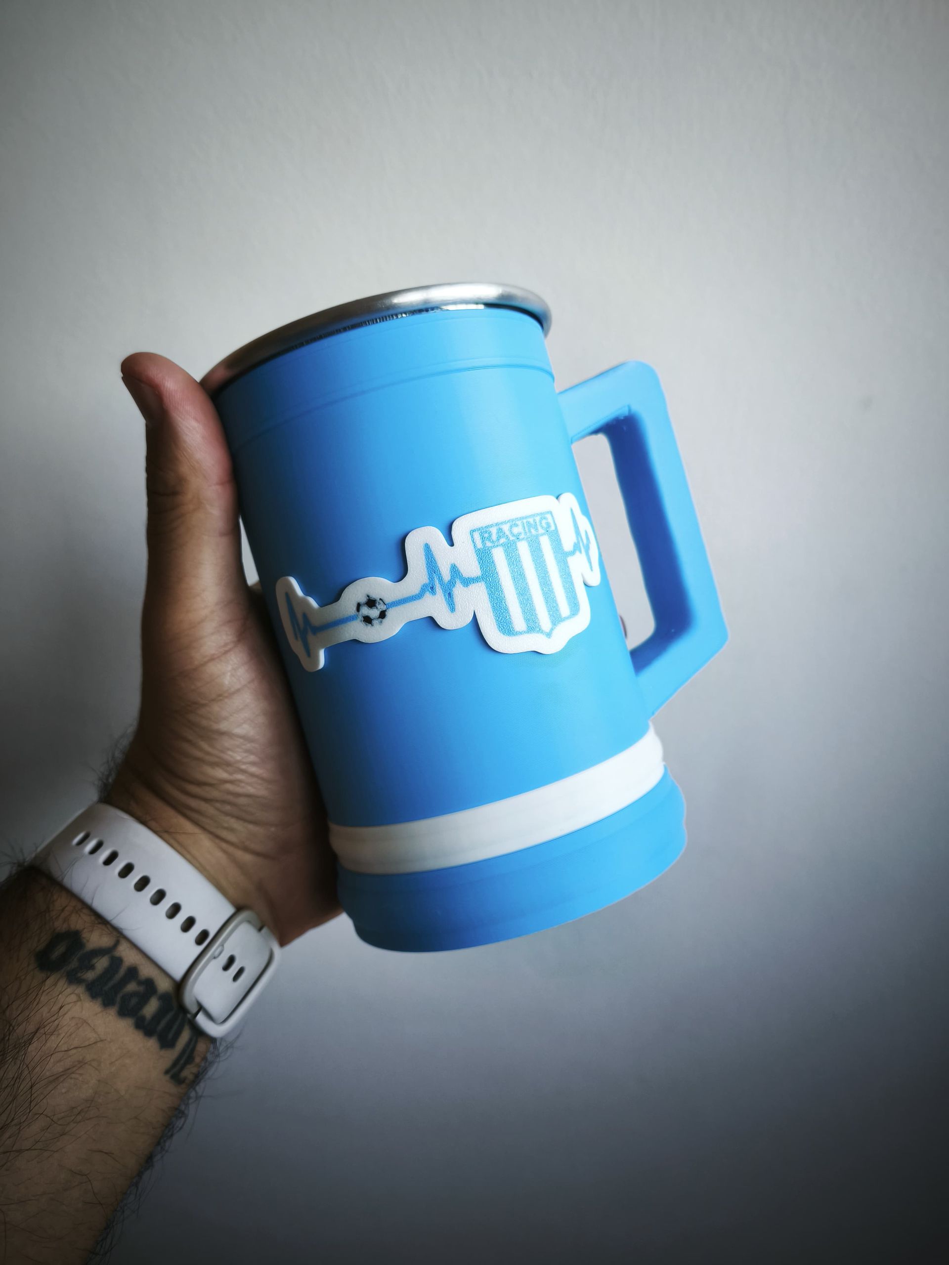 Persona sosteniendo una taza azul claro con un emblema blanco y azul. La taza tiene un borde metálico y una franja blanca.