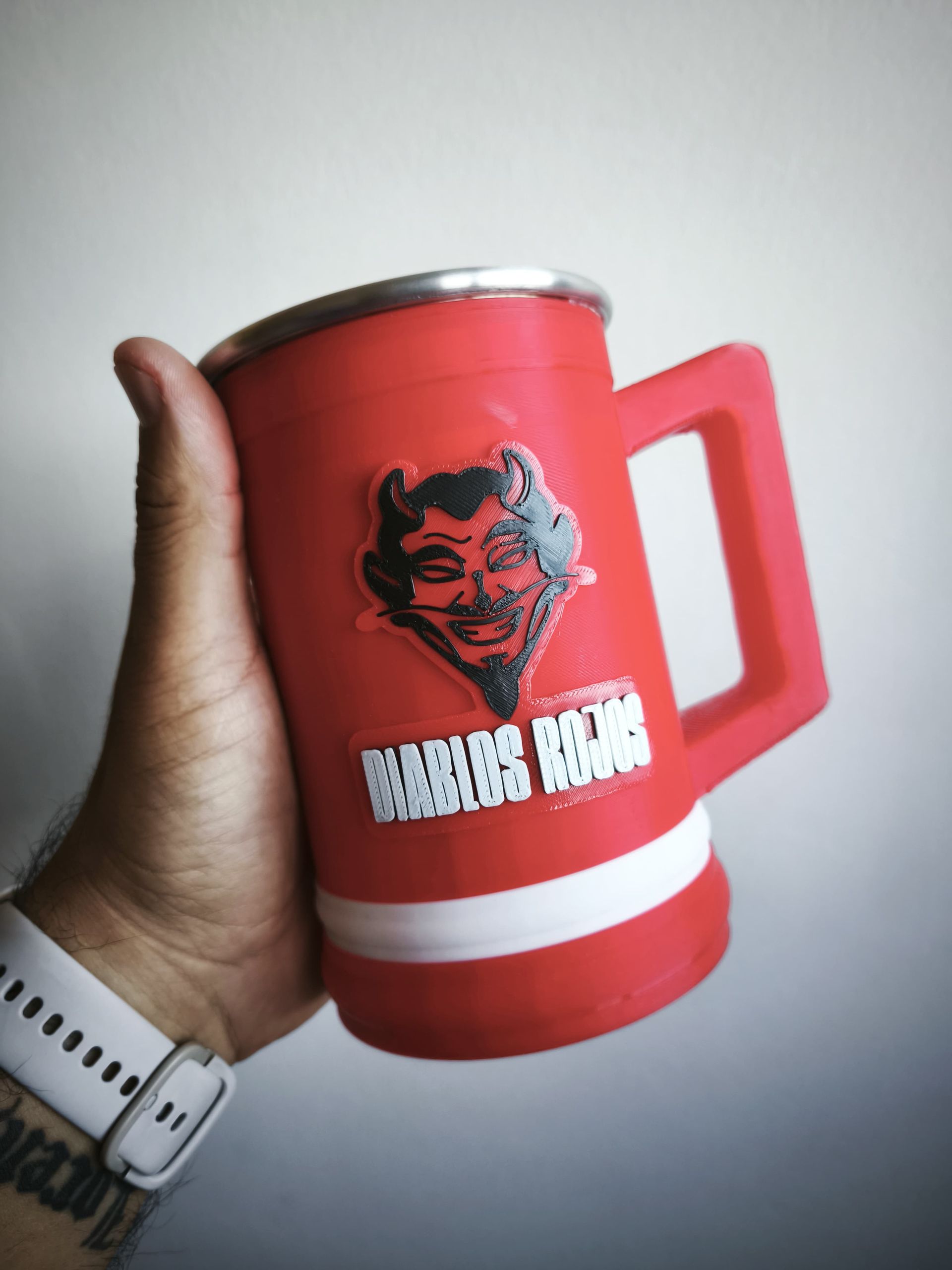 Una mano sosteniendo una taza roja con una raya blanca y el escudo de Argentinos Juniors, sobre un fondo blanco.