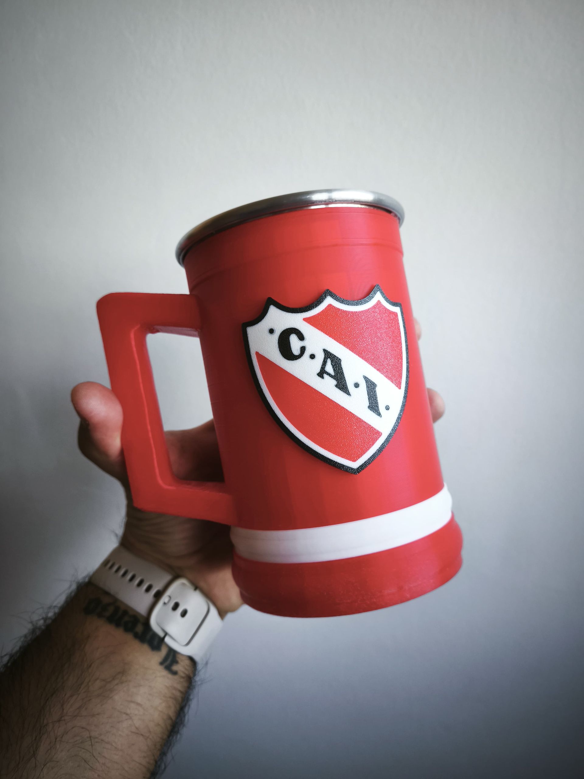 Una mano sosteniendo una taza roja con una raya blanca y el escudo de Argentinos Juniors, sobre un fondo blanco.