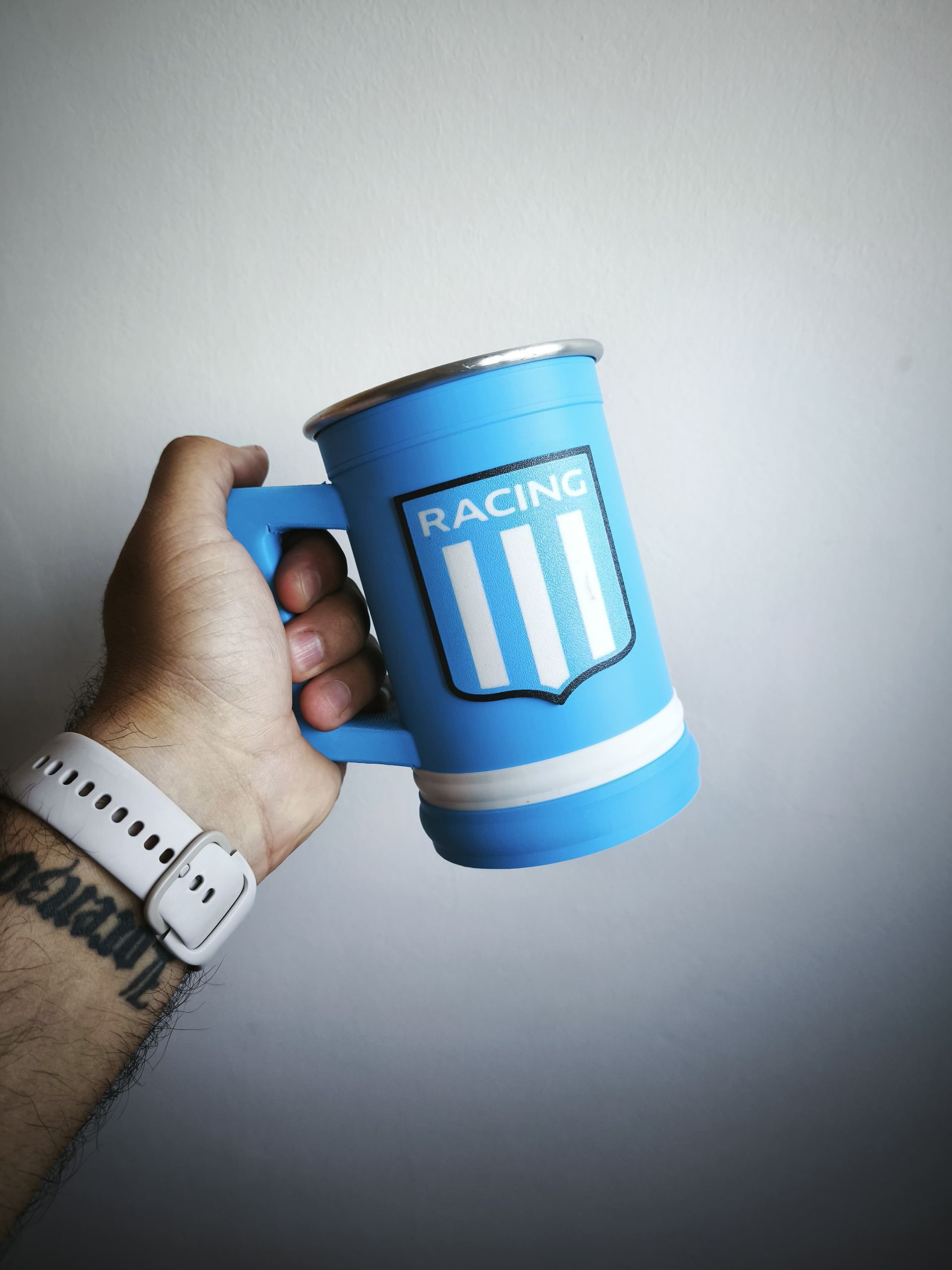 Persona sosteniendo una taza azul claro con un emblema blanco y azul. La taza tiene un borde metálico y una franja blanca.