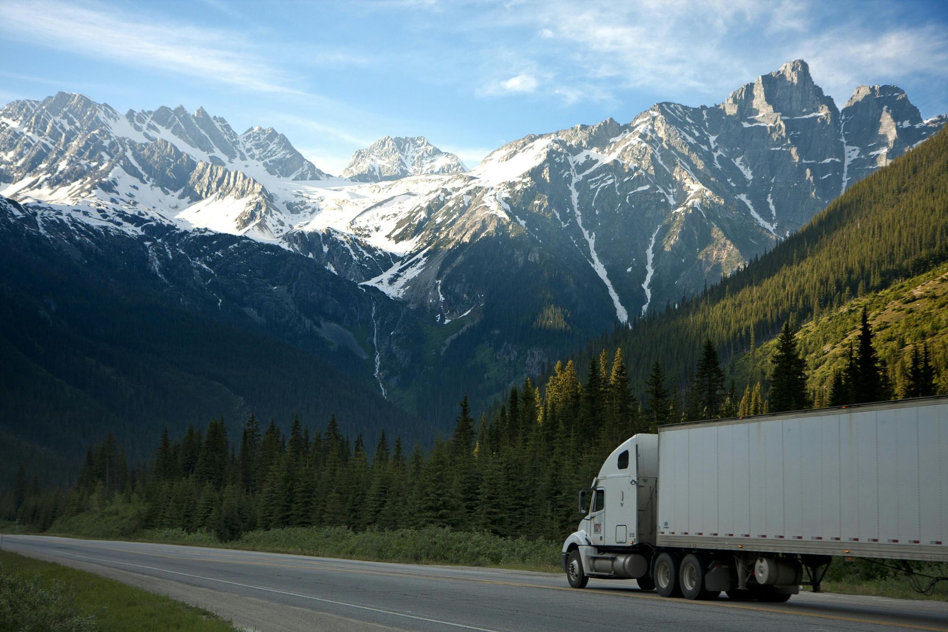 Semi-truck driving on a road winding through a mountain range under a blue sky.