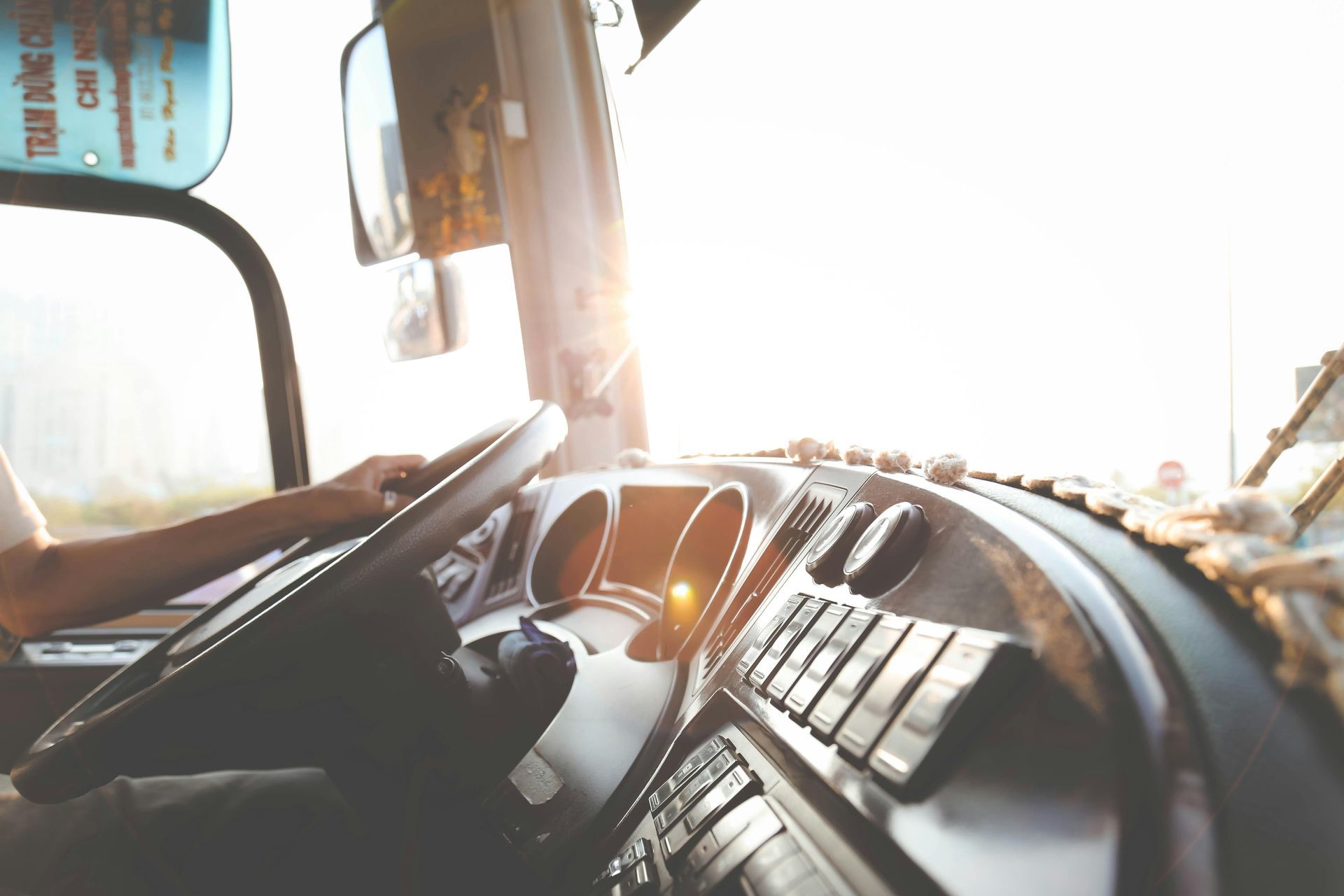 Inside of a bus cockpit, hands gripping the steering wheel, bright sunlight streaming in.