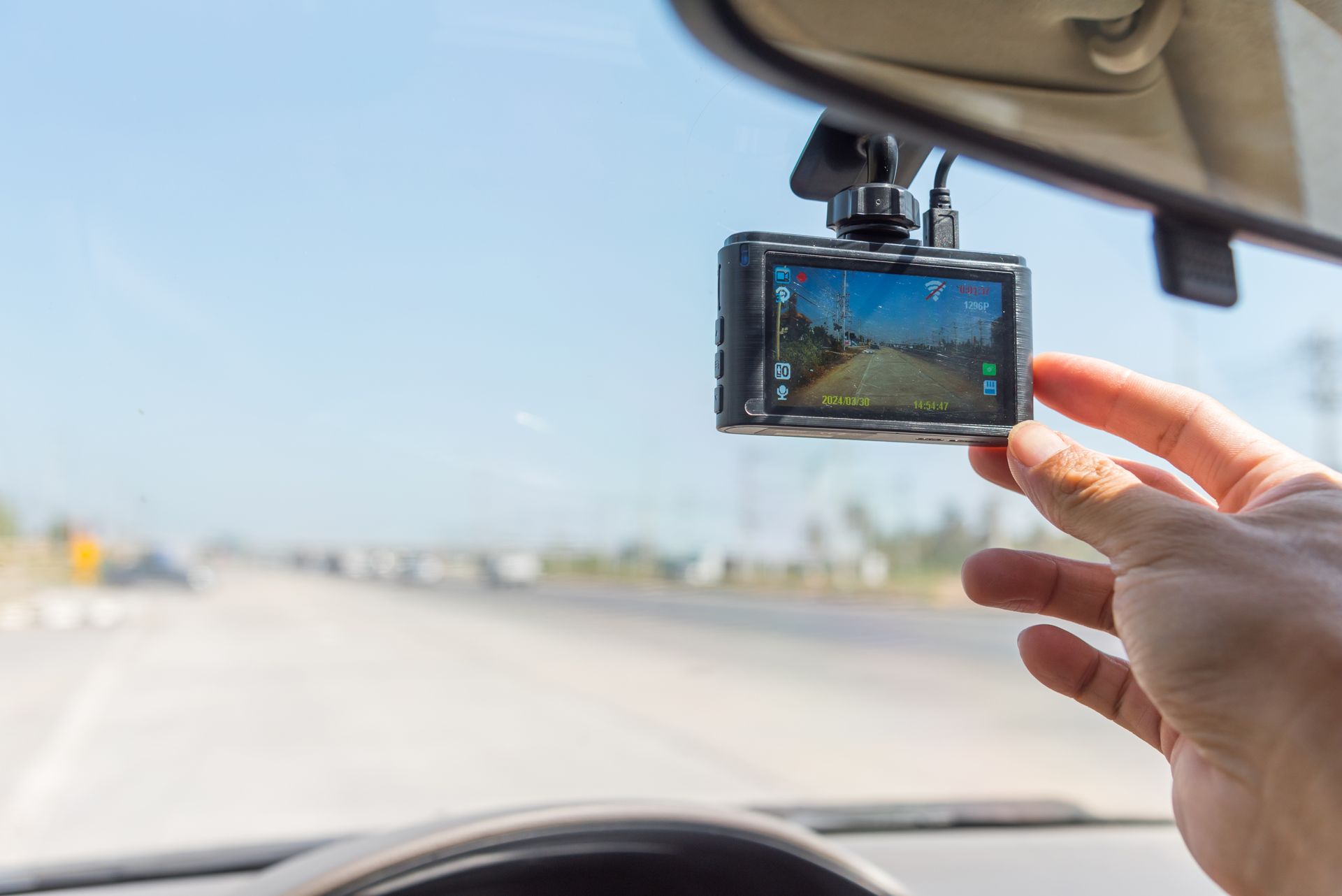 Hand adjusting a dashcam mounted on a car windshield, with a highway visible in the background.