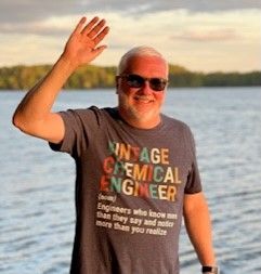 A man wearing a vintage chemical engineer t-shirt is waving at the camera.