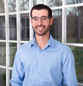 A man wearing glasses and a blue shirt is smiling in front of a window.