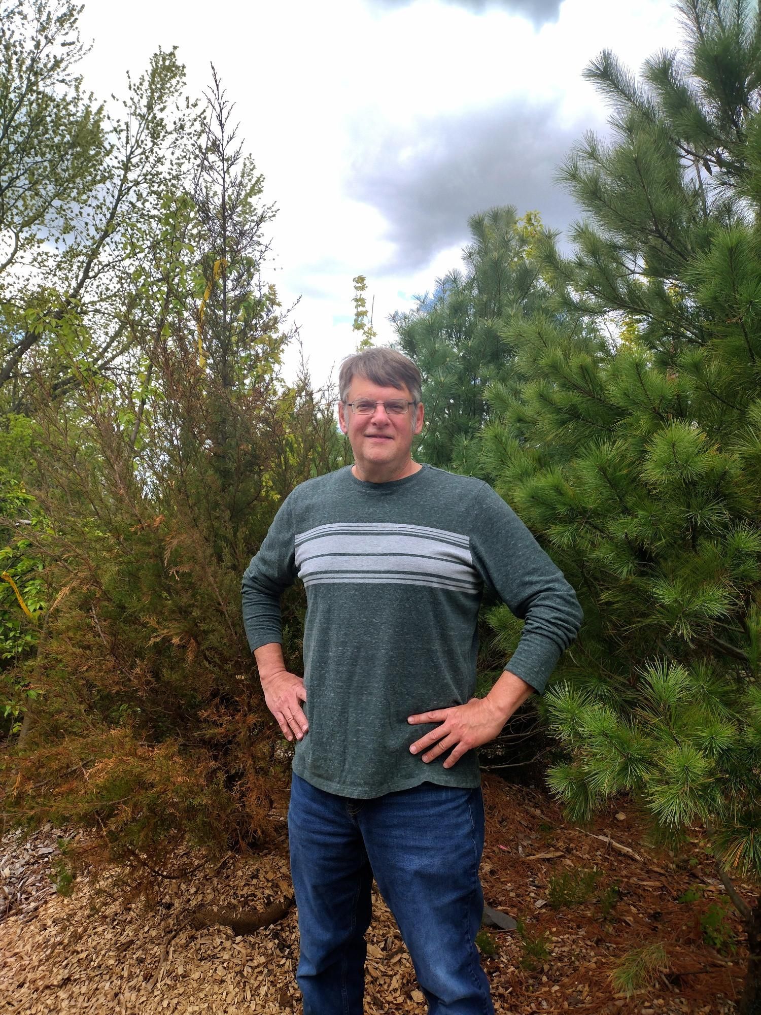 A man is standing in front of a forest with his hands on his hips.