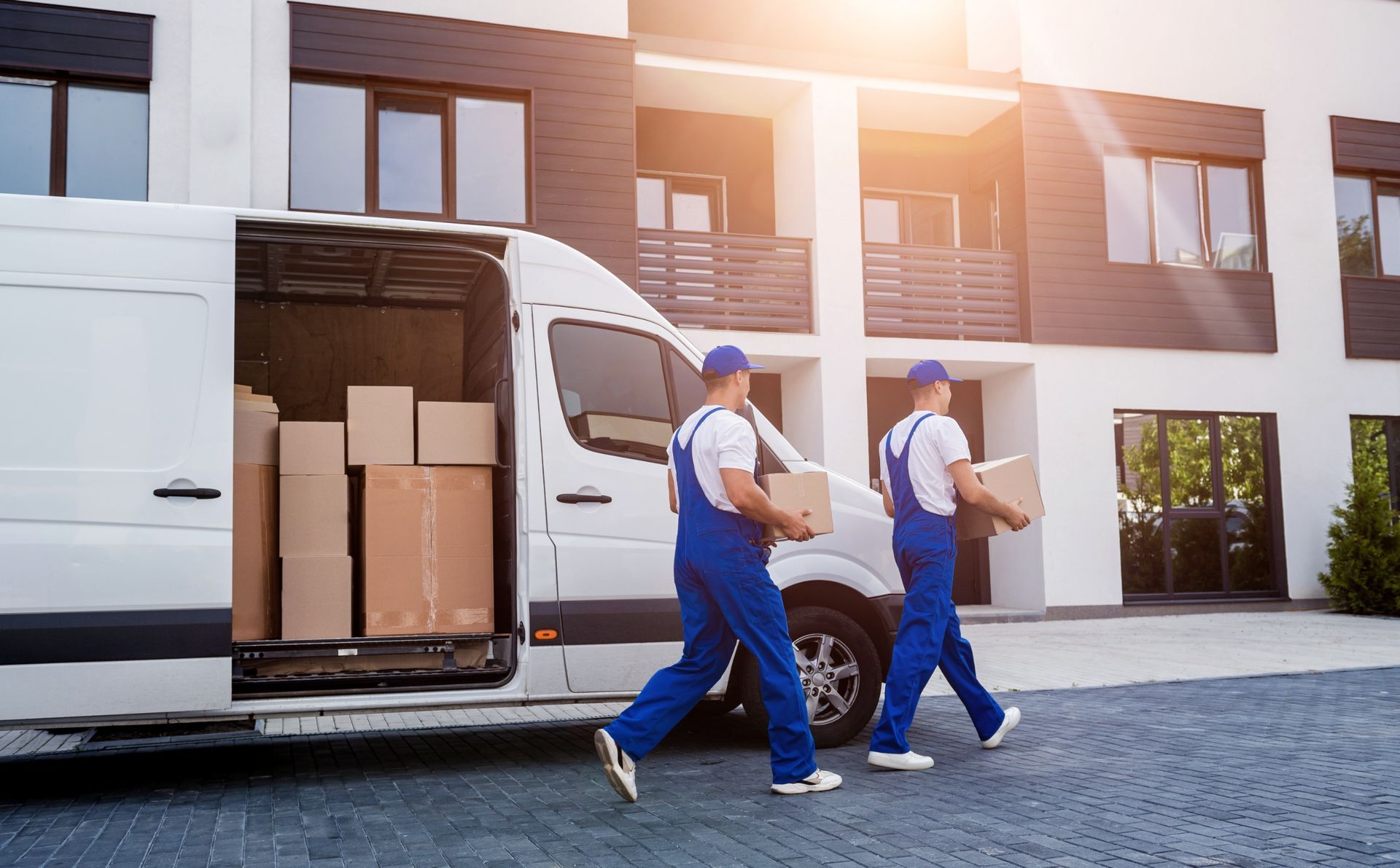 Two workers in blue uniforms carry cardboard boxes from an open white delivery van toward a modern residential building.