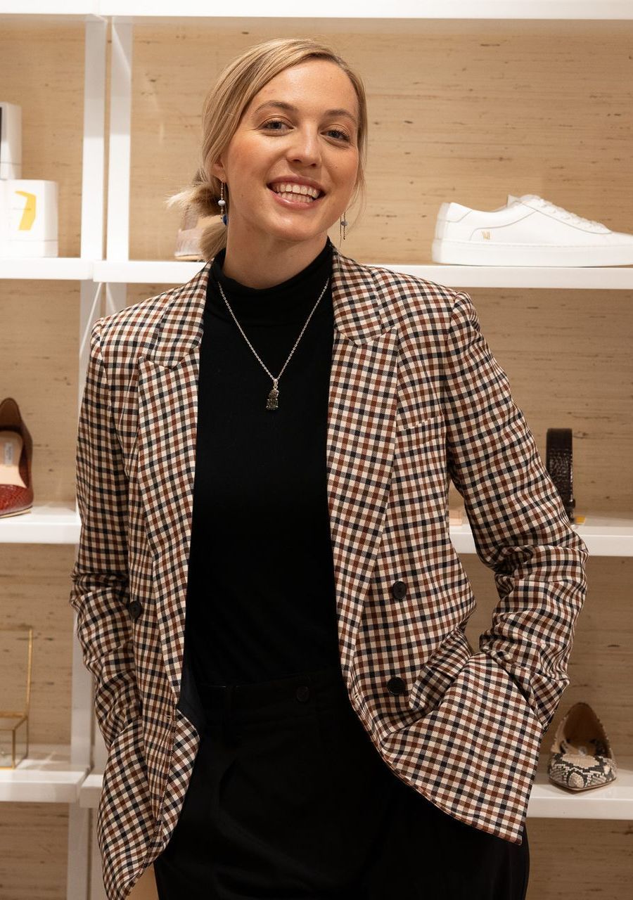 A woman wearing a plaid jacket and a black turtleneck is standing in front of a shoe shelf.