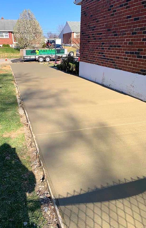 Freshly poured concrete walkway next to a brick building; green grass and a trailer in the background.